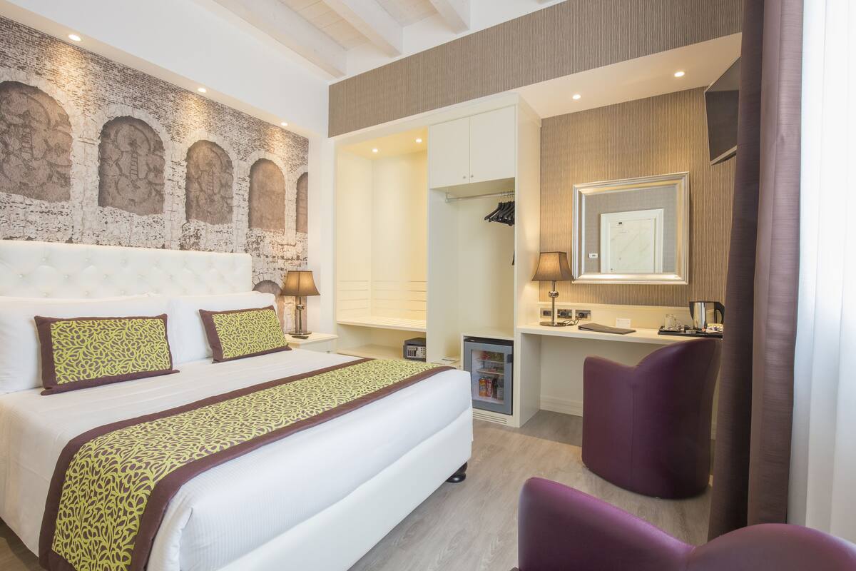 Ein modernes Hotelzimmer mit einem großen Bett, einem Schminktisch mit Spiegel, einer kleinen Kühlbox und einem violetten Sessel.
