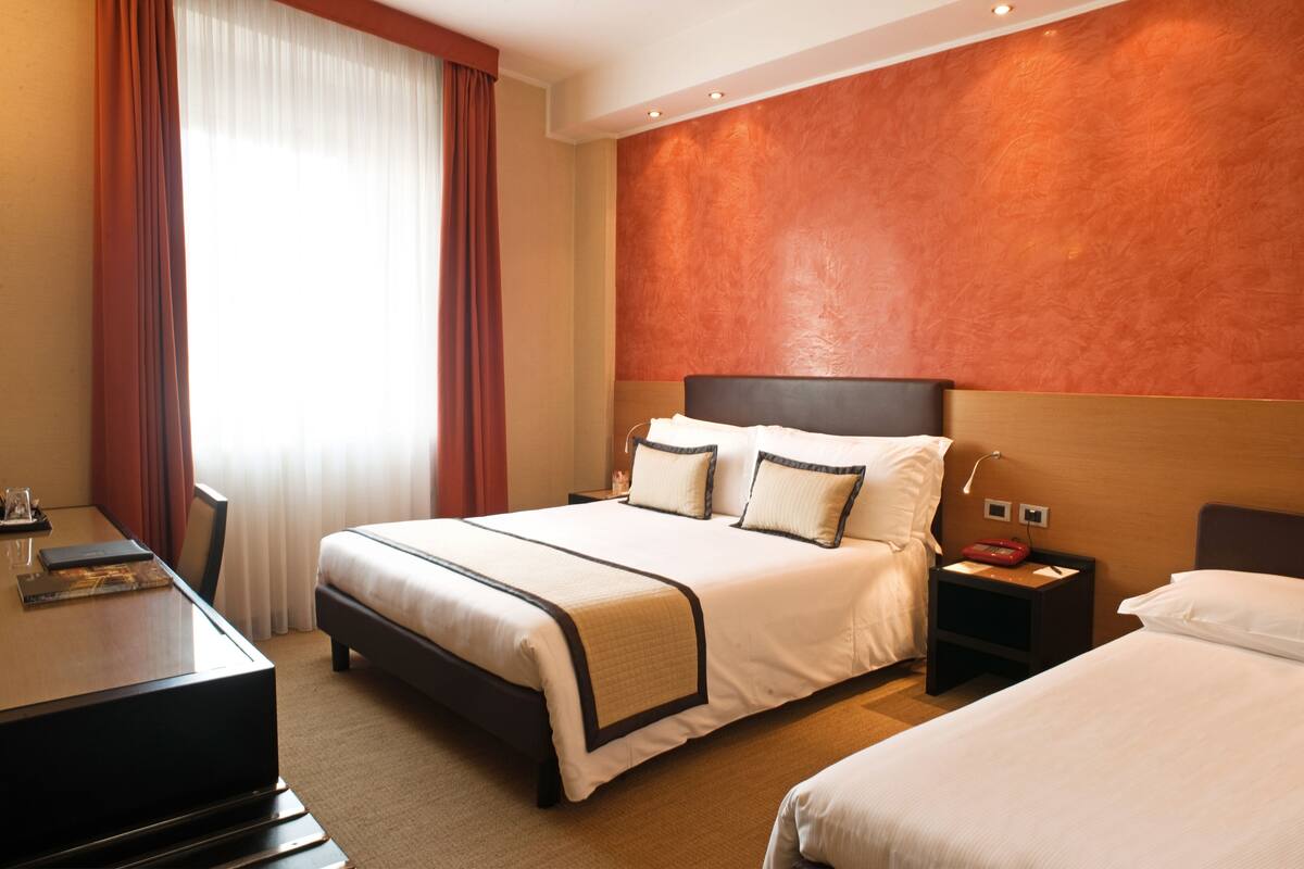 Ein Hotelzimmer mit einem großen Bett, Nachttischen, einem Schreibtisch und einem fenster mit Vorhängen.
