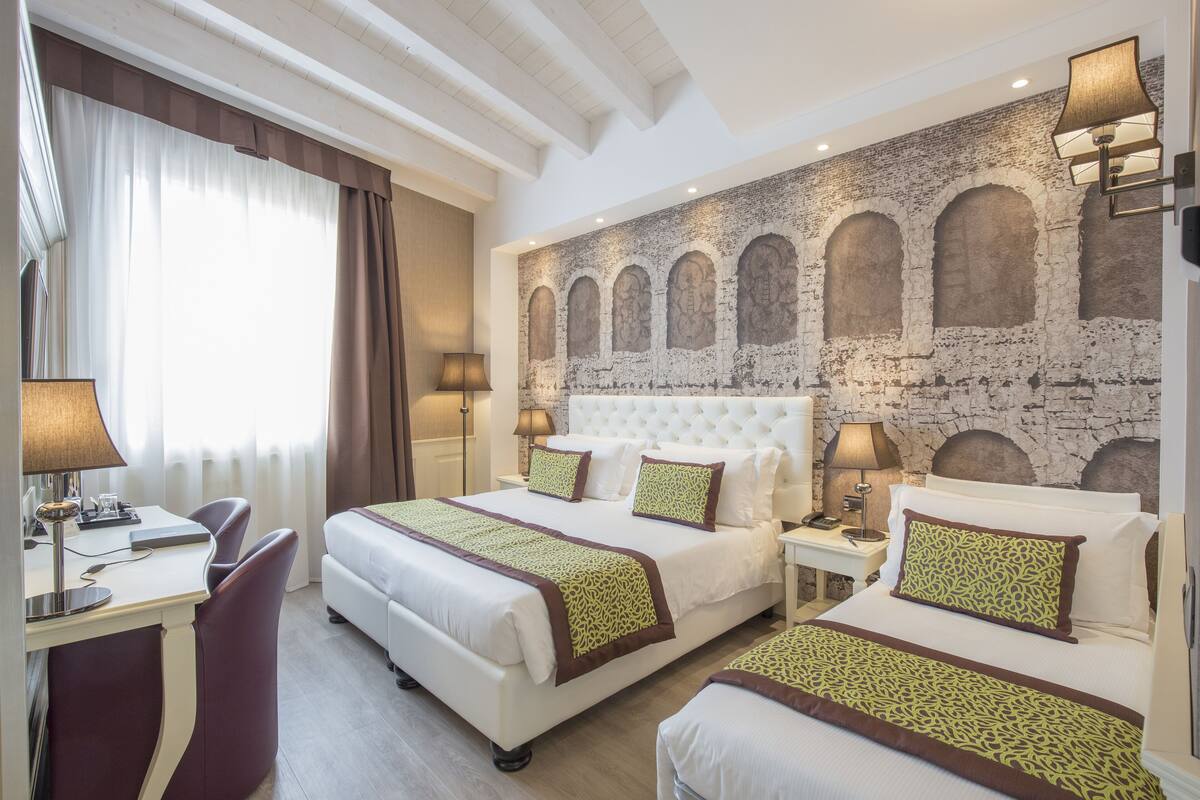Ein Hotelzimmer mit zwei Betten, einem Schreibtisch und einem Stuhl. An der Wand befindet sich ein großes Wandbild mit Bögen und einer strukturierten Oberfläche.