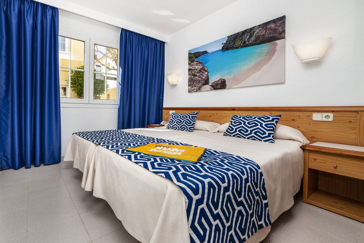 A neatly made bed with a patterned bedspread and pillows, a bedside table with a lamp, and a painting of a beach scene on the wall.