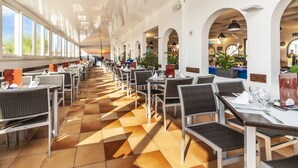 A spacious indoor dining area with high ceilings, large windows, and a checkered floor pattern. Tables and chairs are neatly arranged, and potted plants are placed around the room. - Apartamentos Globales Binimar (Ciutadella de Menorca)