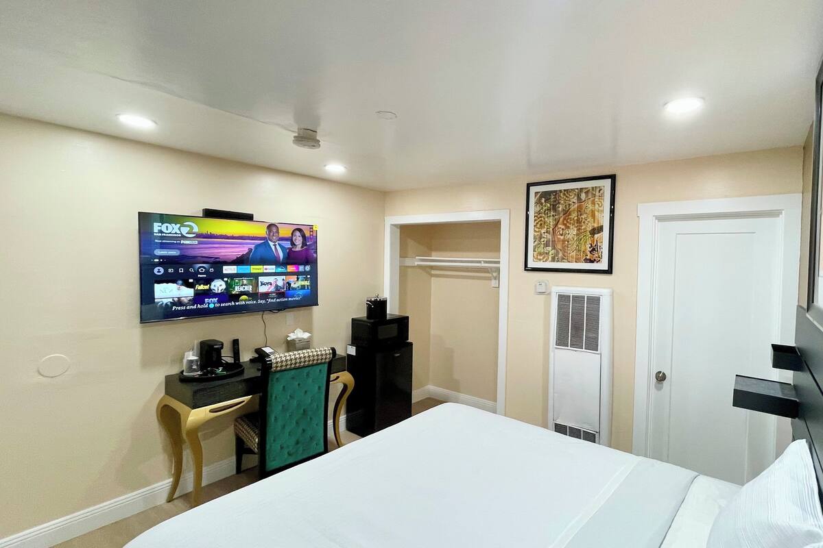A hotel room with a bed, a desk with a computer, a TV mounted on the wall, a door, and a window.