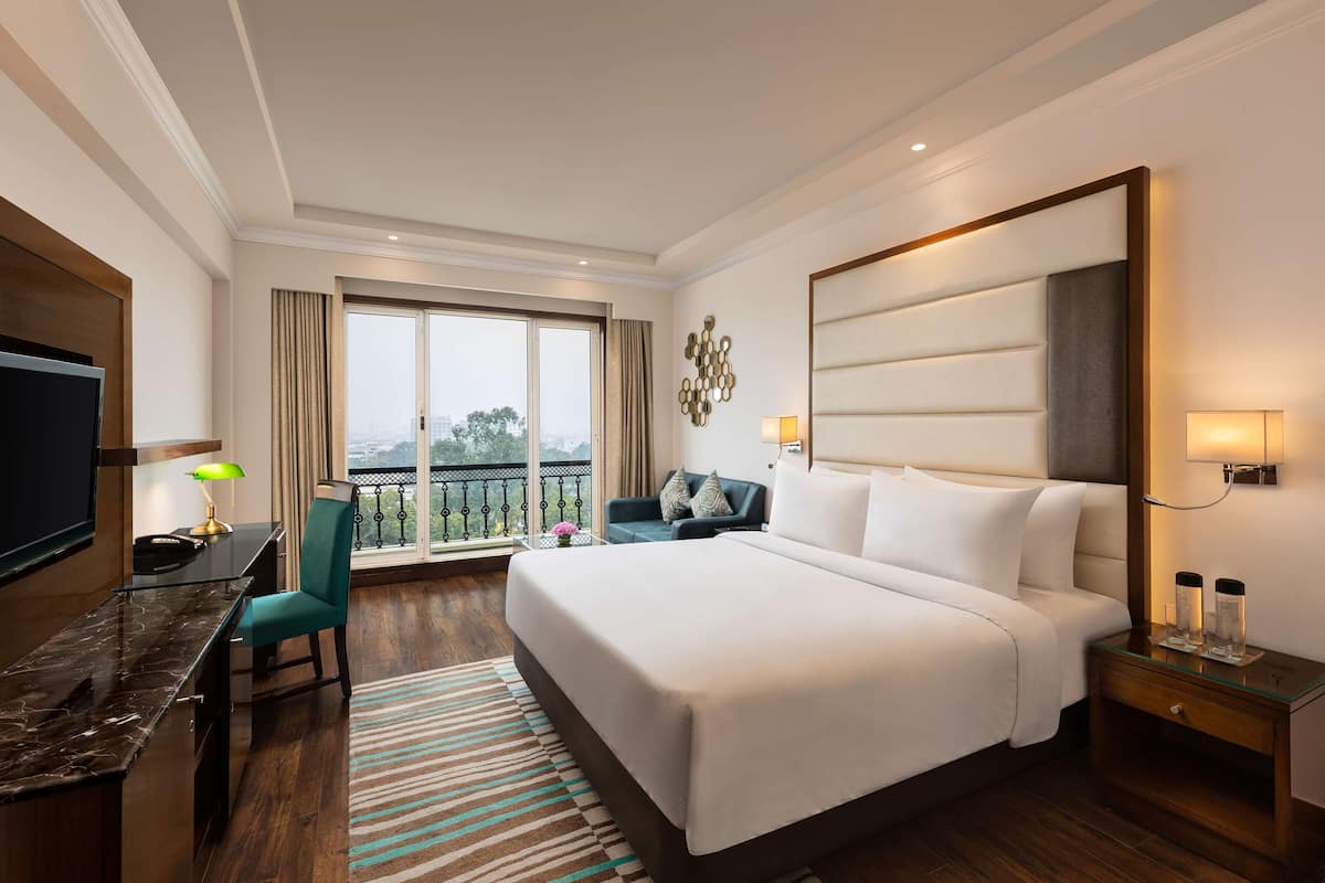 A hotel room with a large bed, a desk with a chair, a TV, and a balcony with a view.