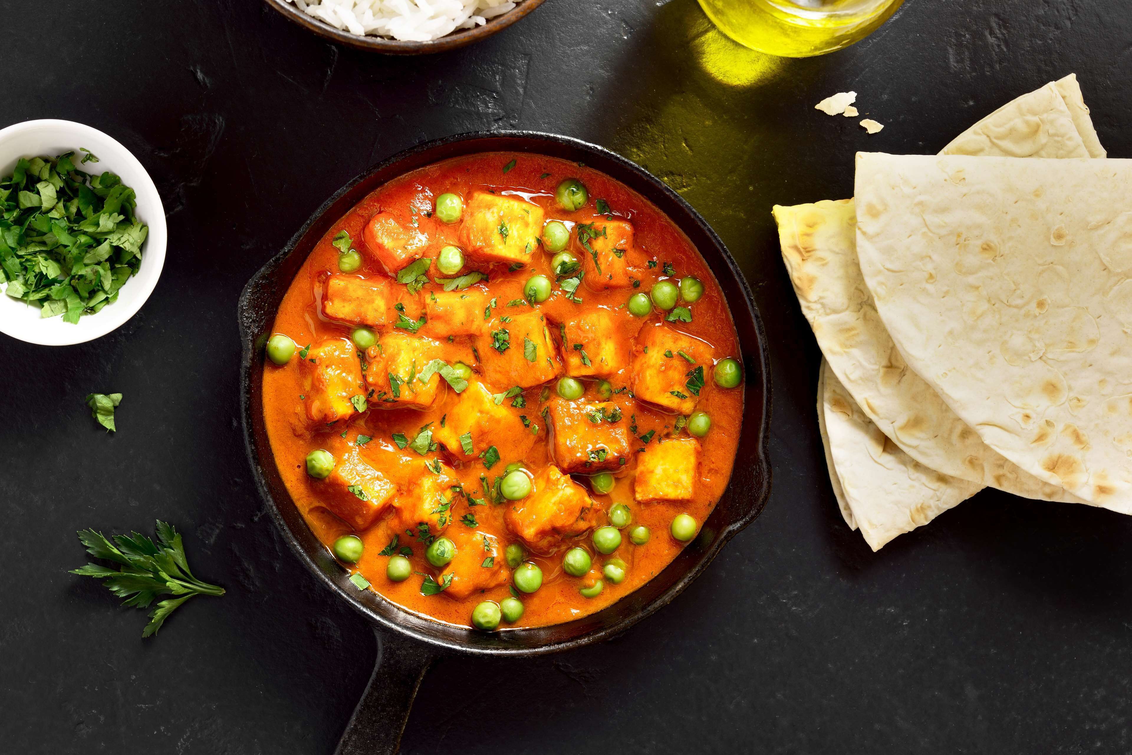A bowl of paneer curry with peas and herbs, accompanied by flatbreads and a small bowl of chopped herbs.
