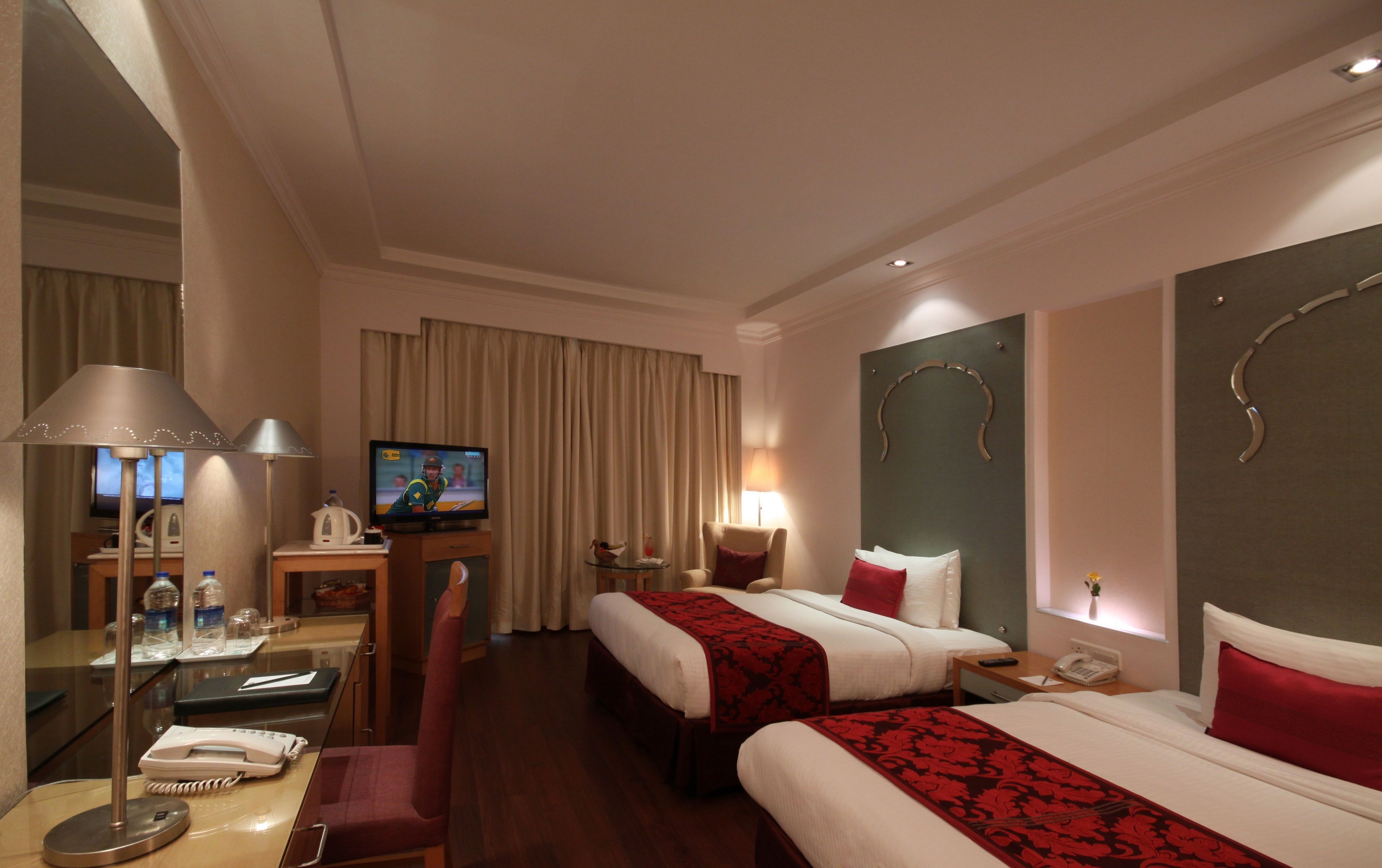 A hotel room with two beds, a desk with a computer, a chair, a TV, and a lamp.