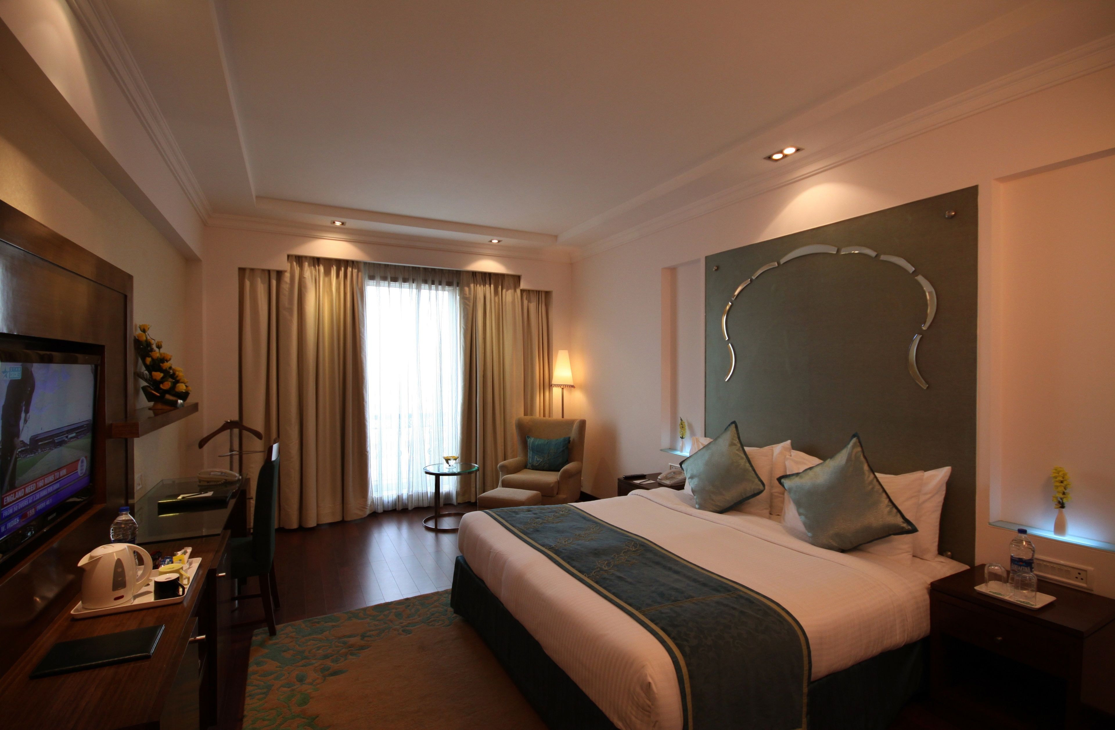 Habitación de hotel con una cama grande, televisión, escritorio con silla, una mesita y ventana con cortinas.