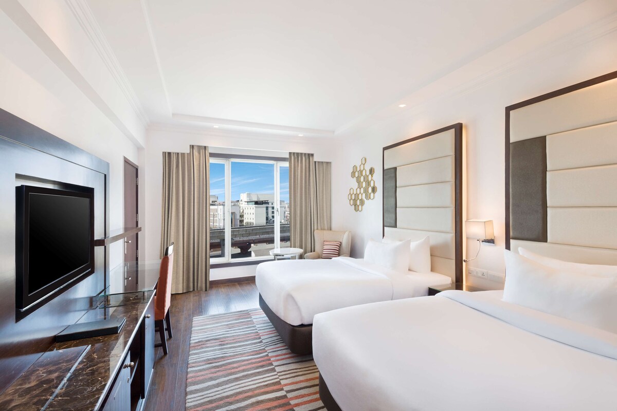 A hotel room with two beds, a TV, a desk, and a view of the city.