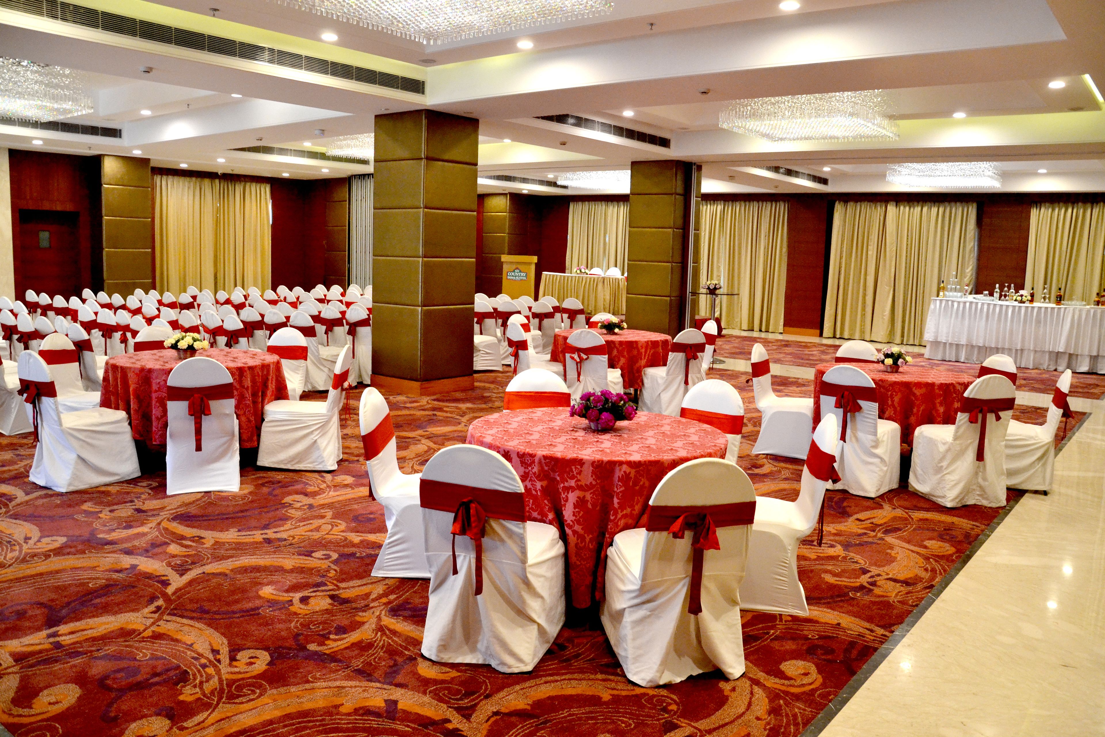 Uma sala de banquetes com mesas redondas e cadeiras brancas, toalhas de mesa vermelhas e cortinas douradas.