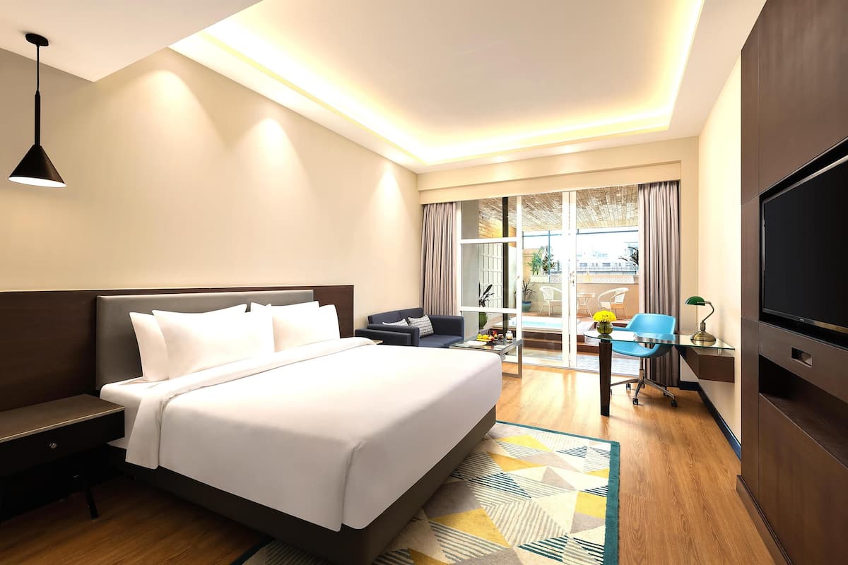 A hotel room with a large bed, a desk with a computer, a TV, and a view of the outside through a large window.