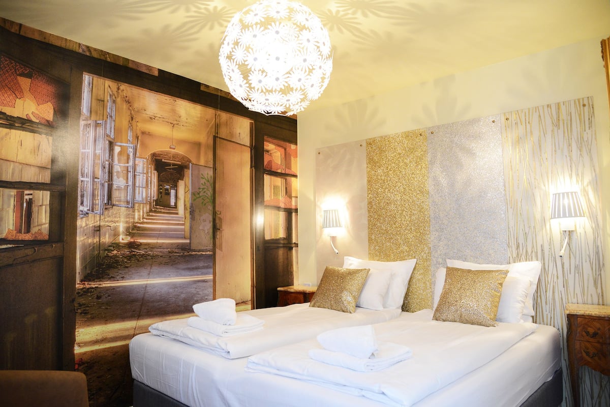 Habitación de hotel con una cama grande, dos mesitas de noche, una araña y un mural en la pared.