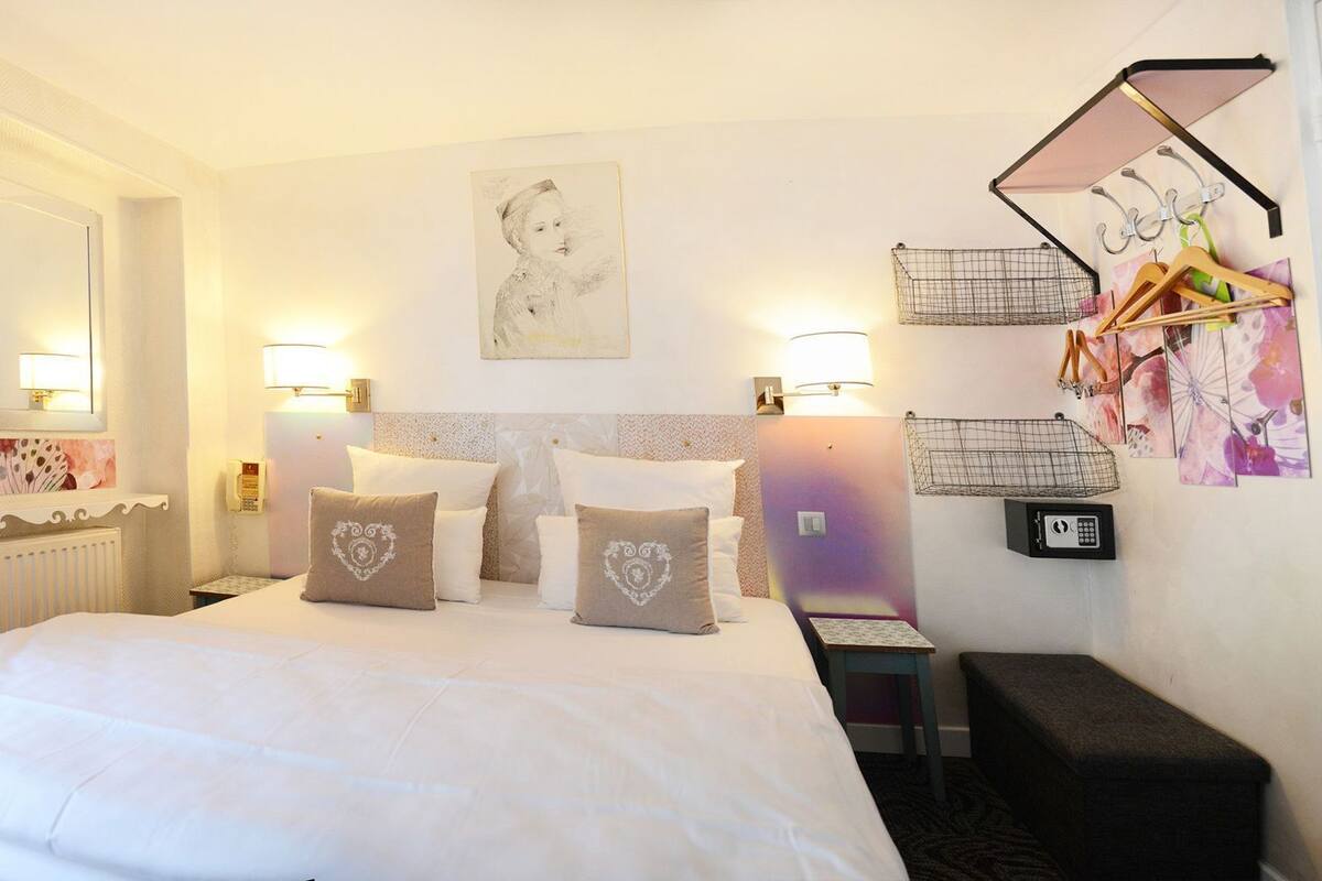 Habitación de hotel con una cama grande, lámparas de noche, cabecero, mesita de noche pequeña, perchero de pared y radiador.