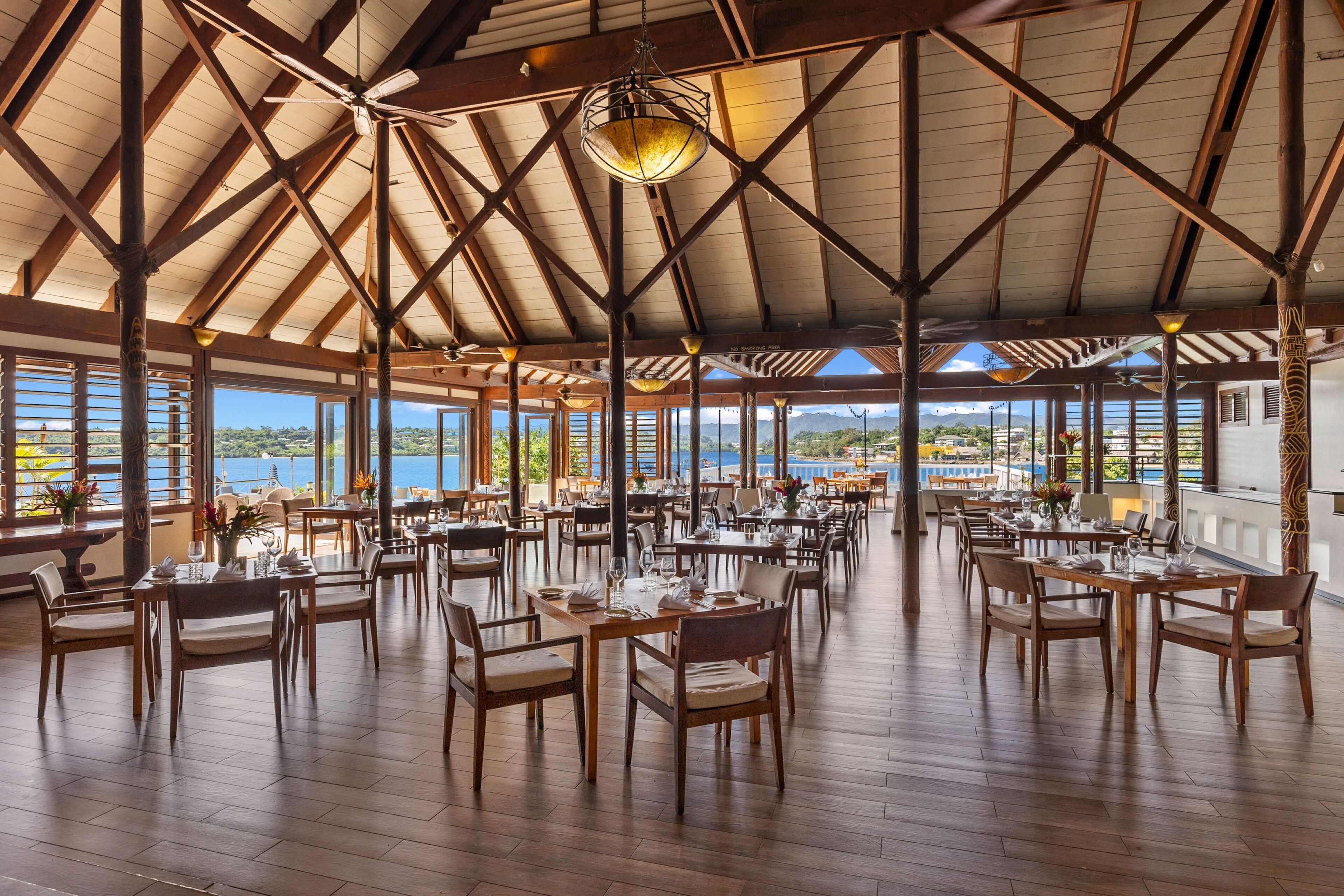 A spacious dining area with wooden tables and chairs, large windows offering views of the sea, and a high ceiling with exposed beams.