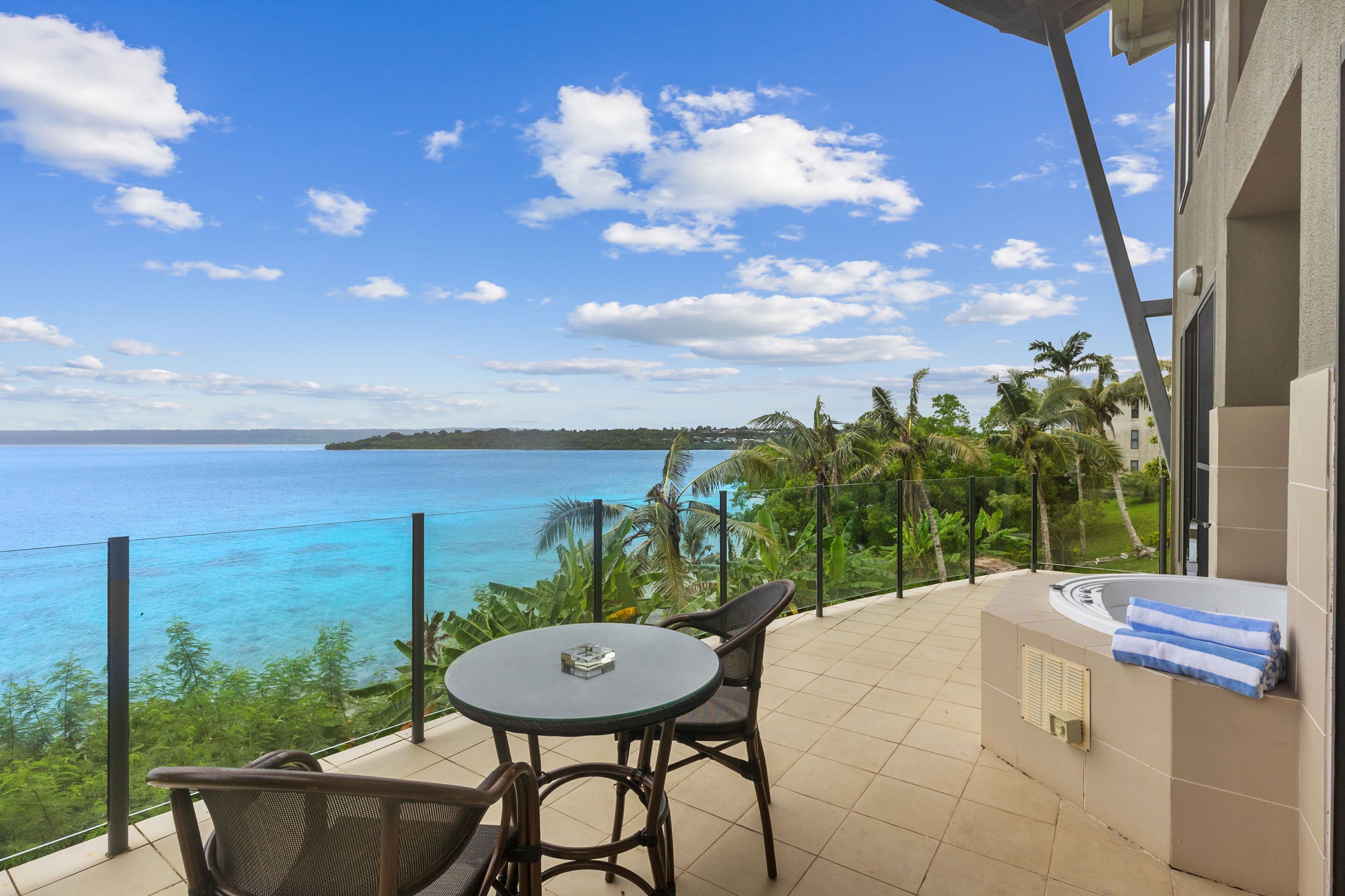 A balcony with a table and chairs overlooking a turquoise sea and palm trees.