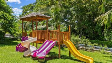 Children can play on the wooden structure with slides in a lush park setting surrounded by trees and well-maintained grass.