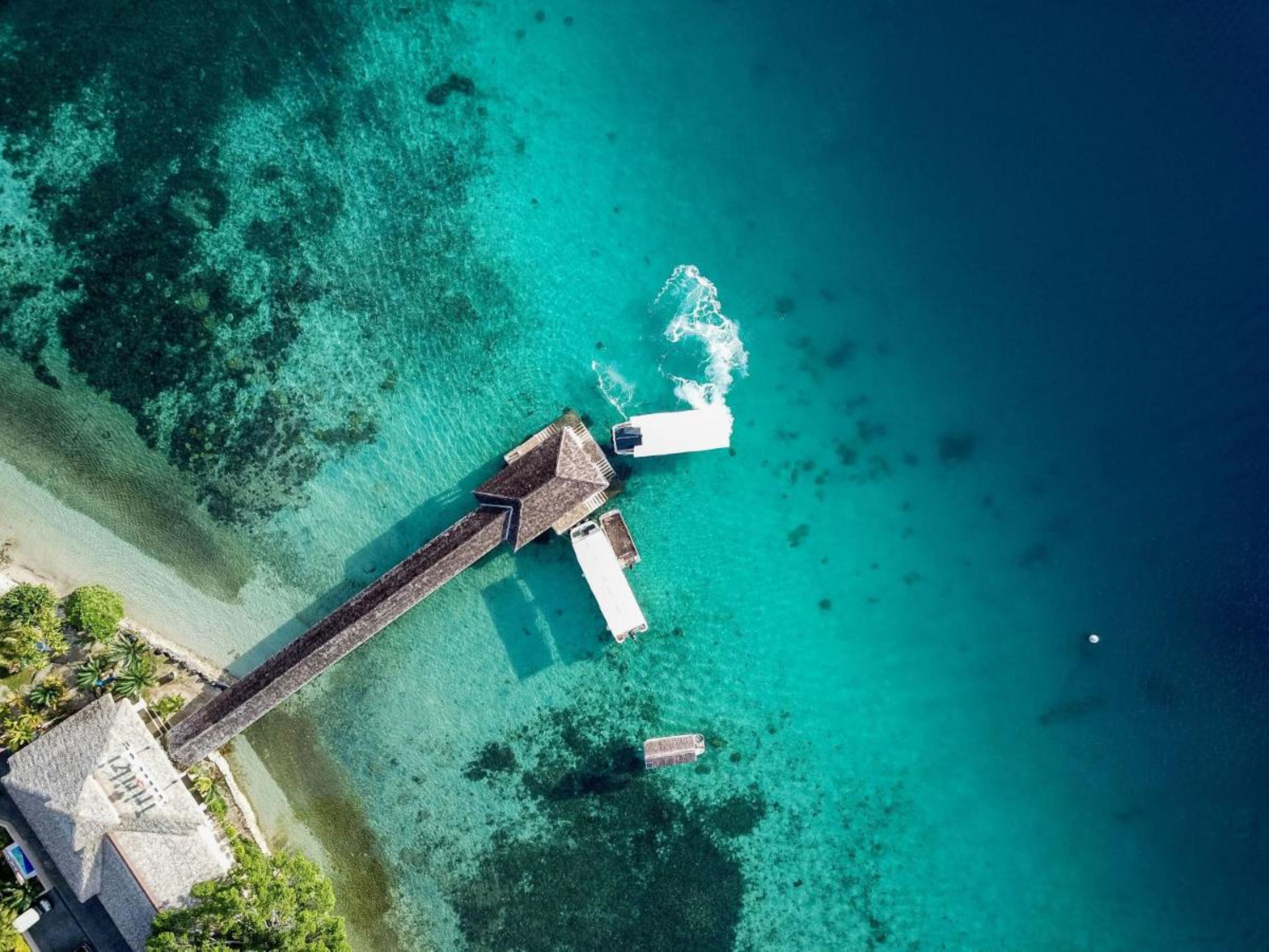 Una vista aerea di una zona costiera con un lungo molo in legno che si protende in un mare color turchese, un piccolo edificio con tetto di paglia vicino alla riva, e una visuale chiara dell'oceano.