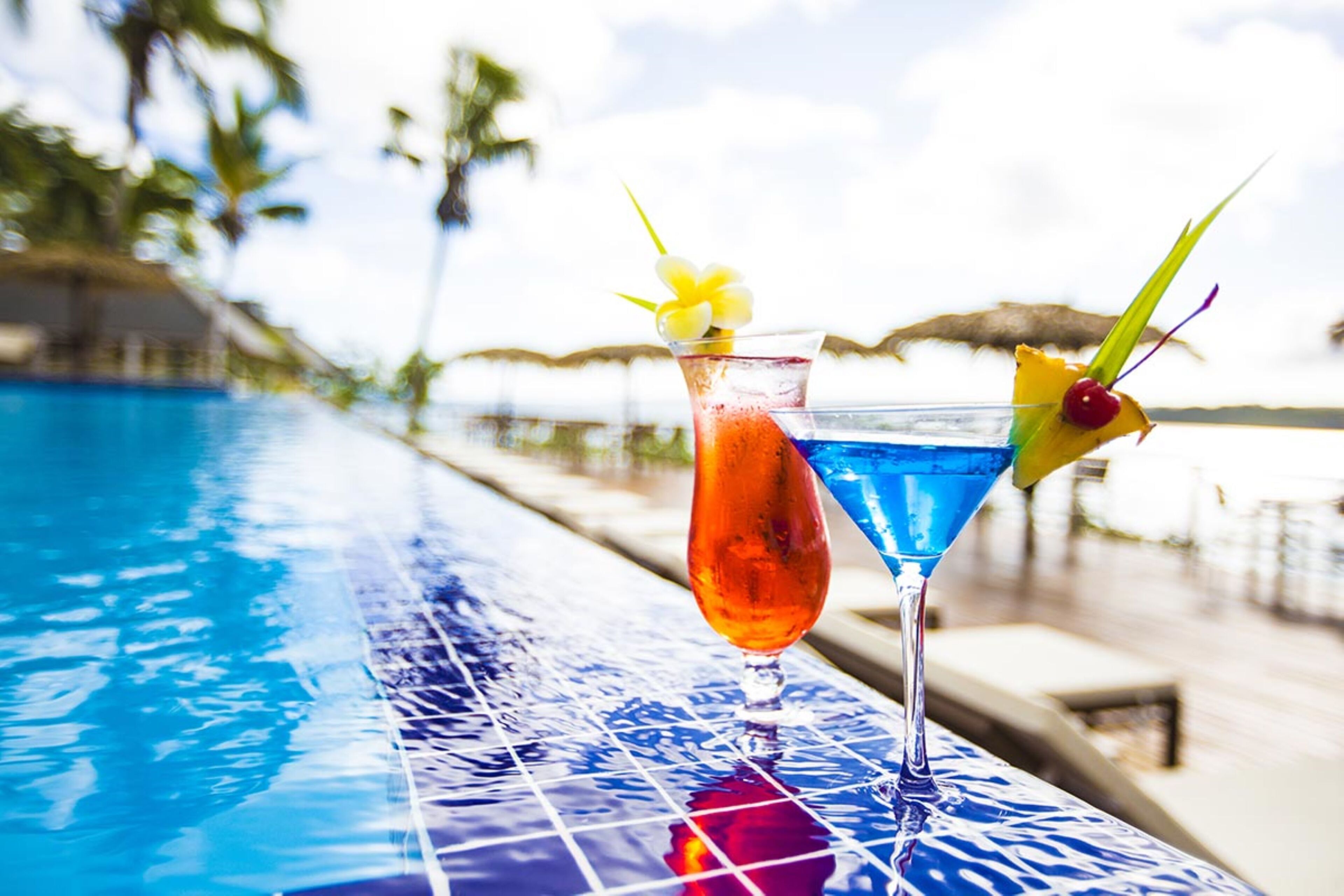 Two cocktails on a poolside table with a clear view of the pool and palm trees in the background.