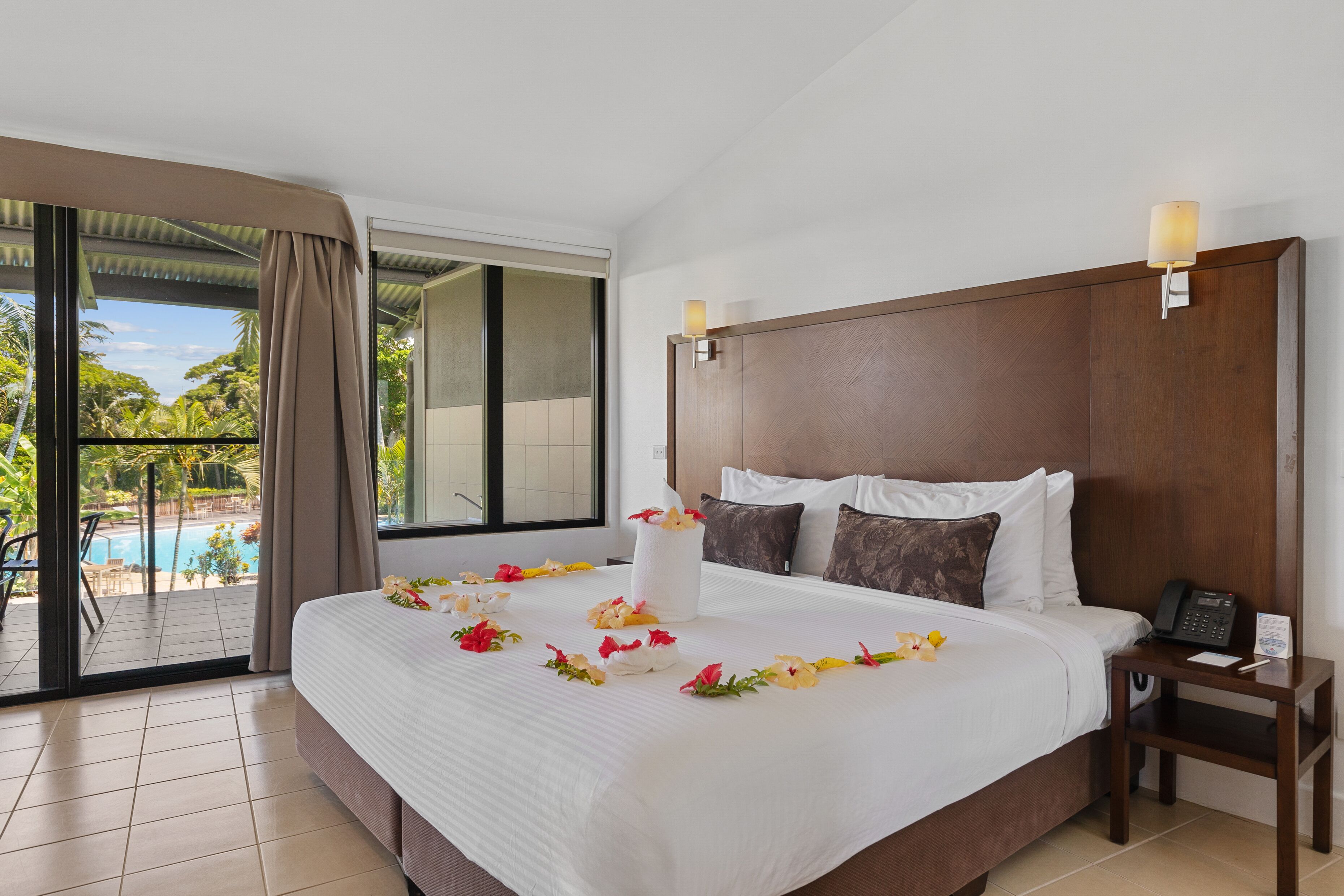 A hotel room with a large bed, a bedside table, a telephone, and a view of a swimming pool.