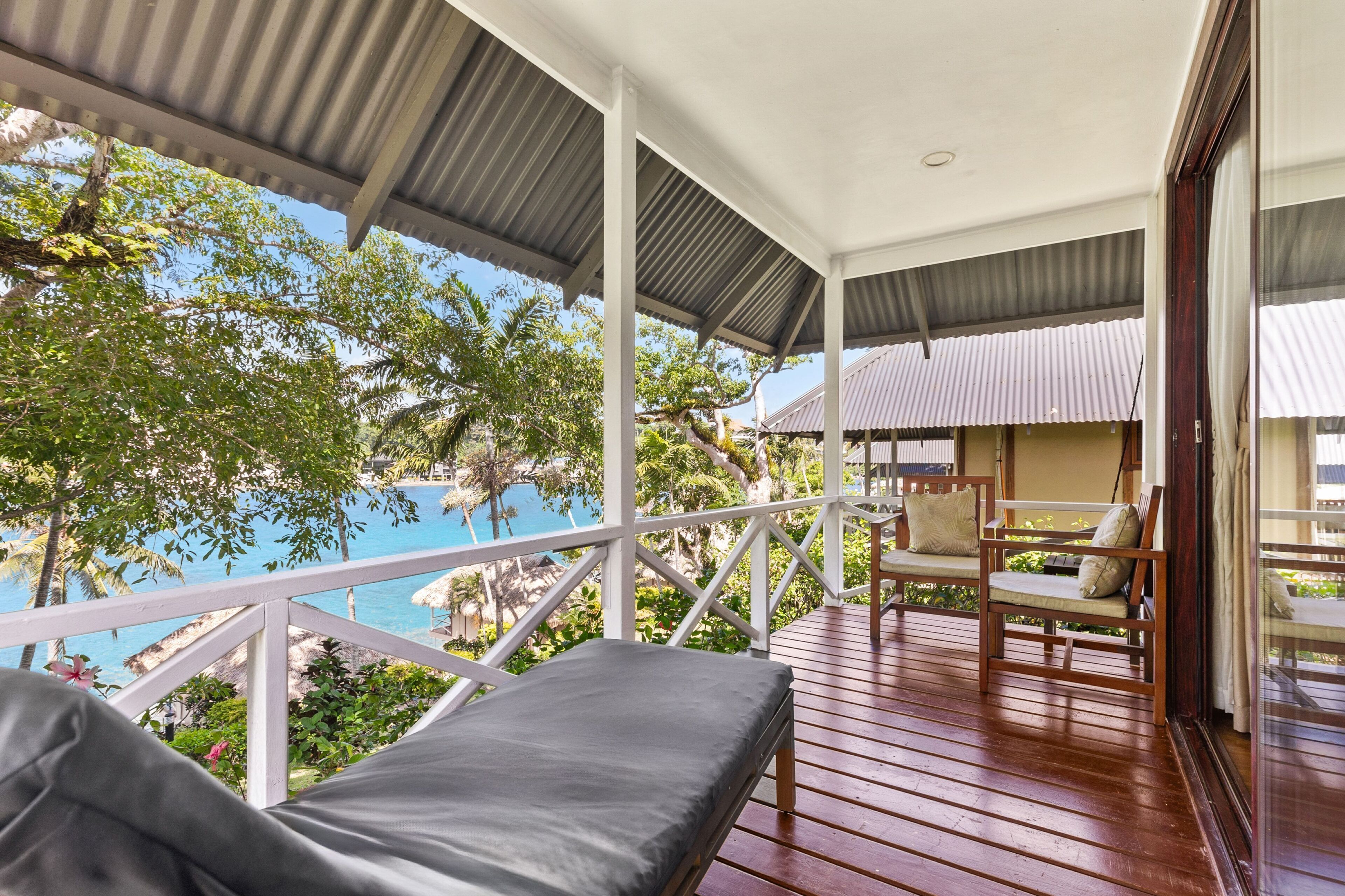 A balcony with wooden flooring, a cushioned chair, and a view of a pool and tropical trees.