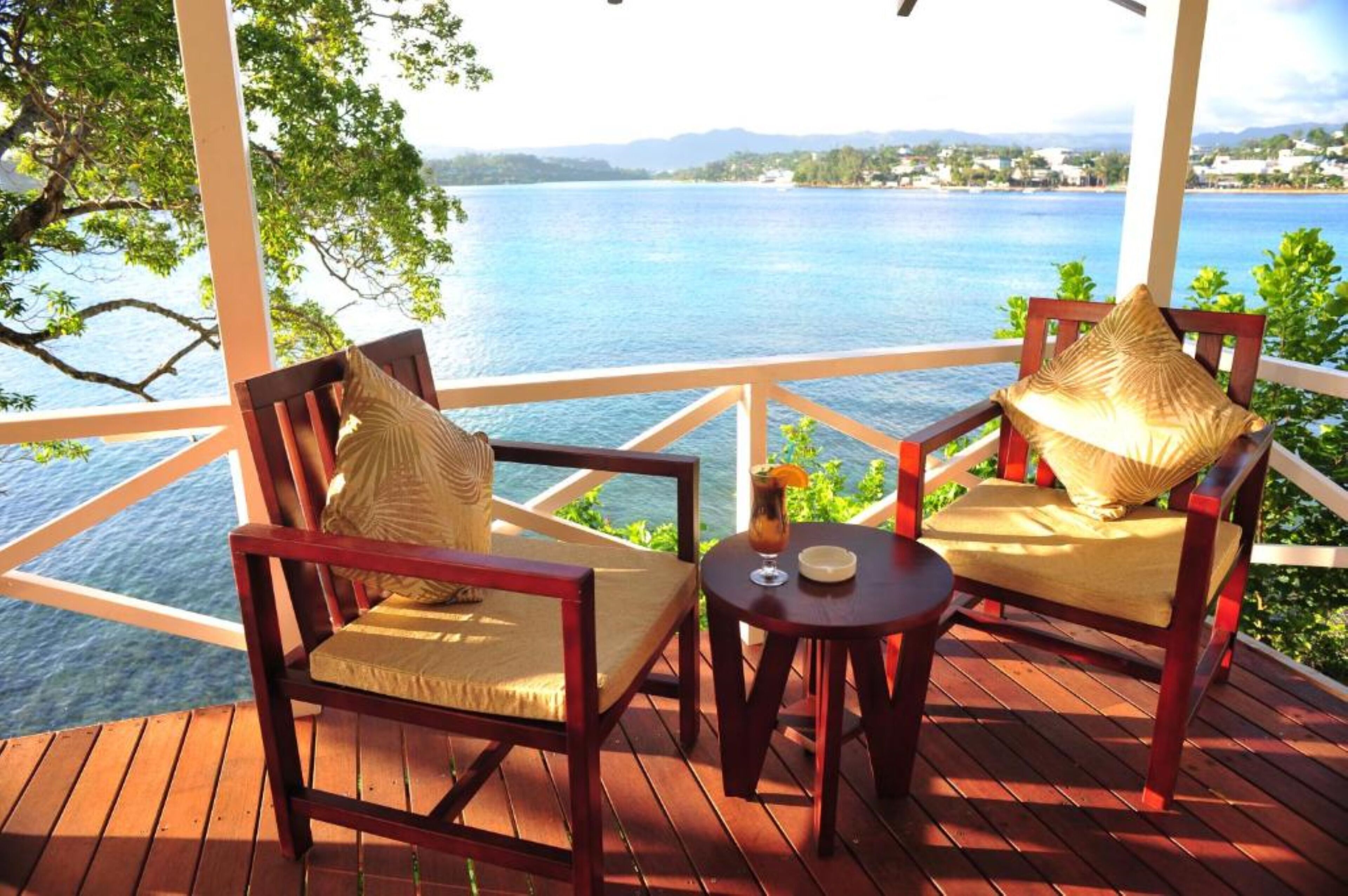 Una terraza de madera con dos sillas de madera y una pequeña mesa, con vistas a una extensa masa de agua y una costa lejana.