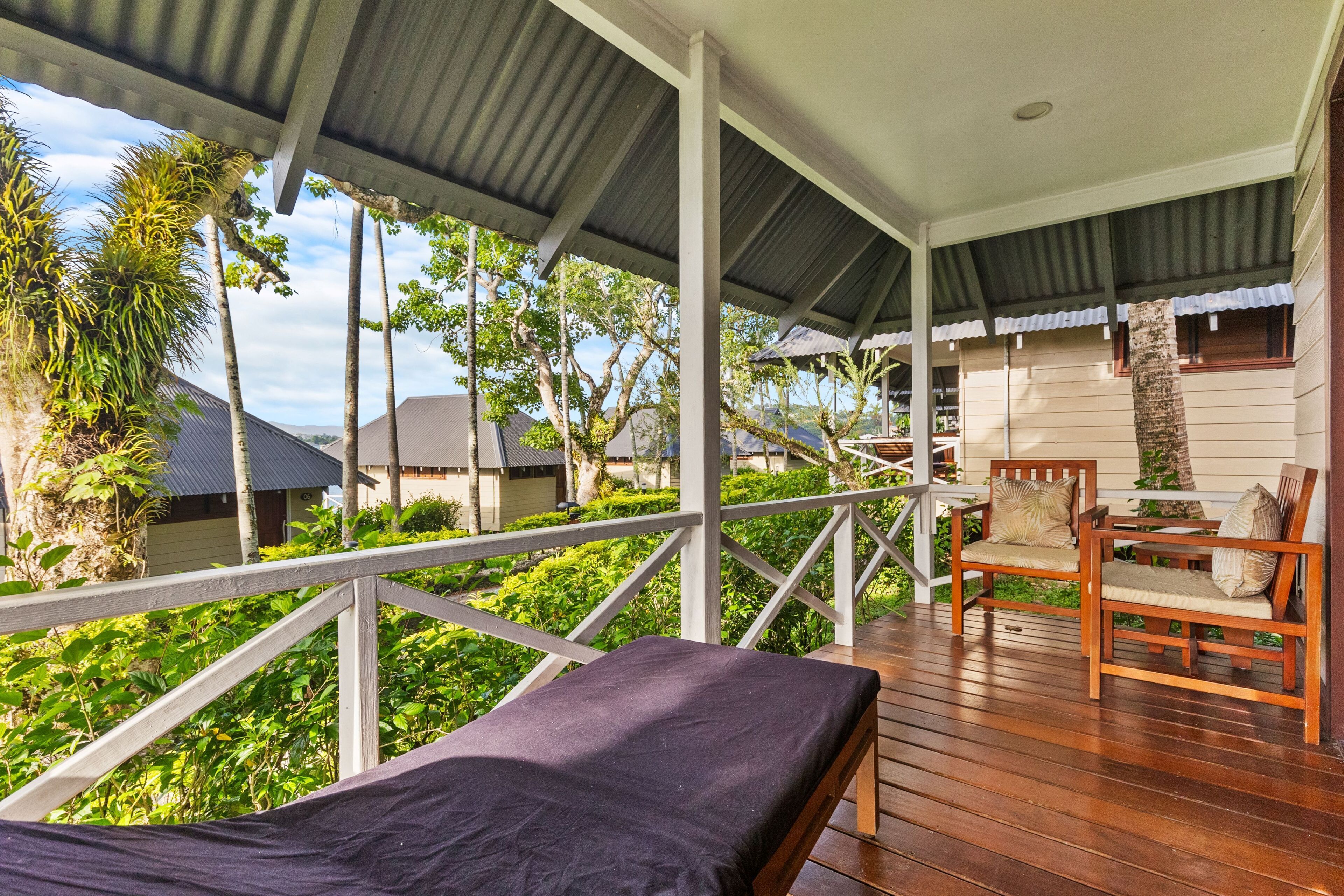 A wooden deck with a bed, two wooden chairs with cushions, and a railing overlooking a garden with trees and a building in the background.