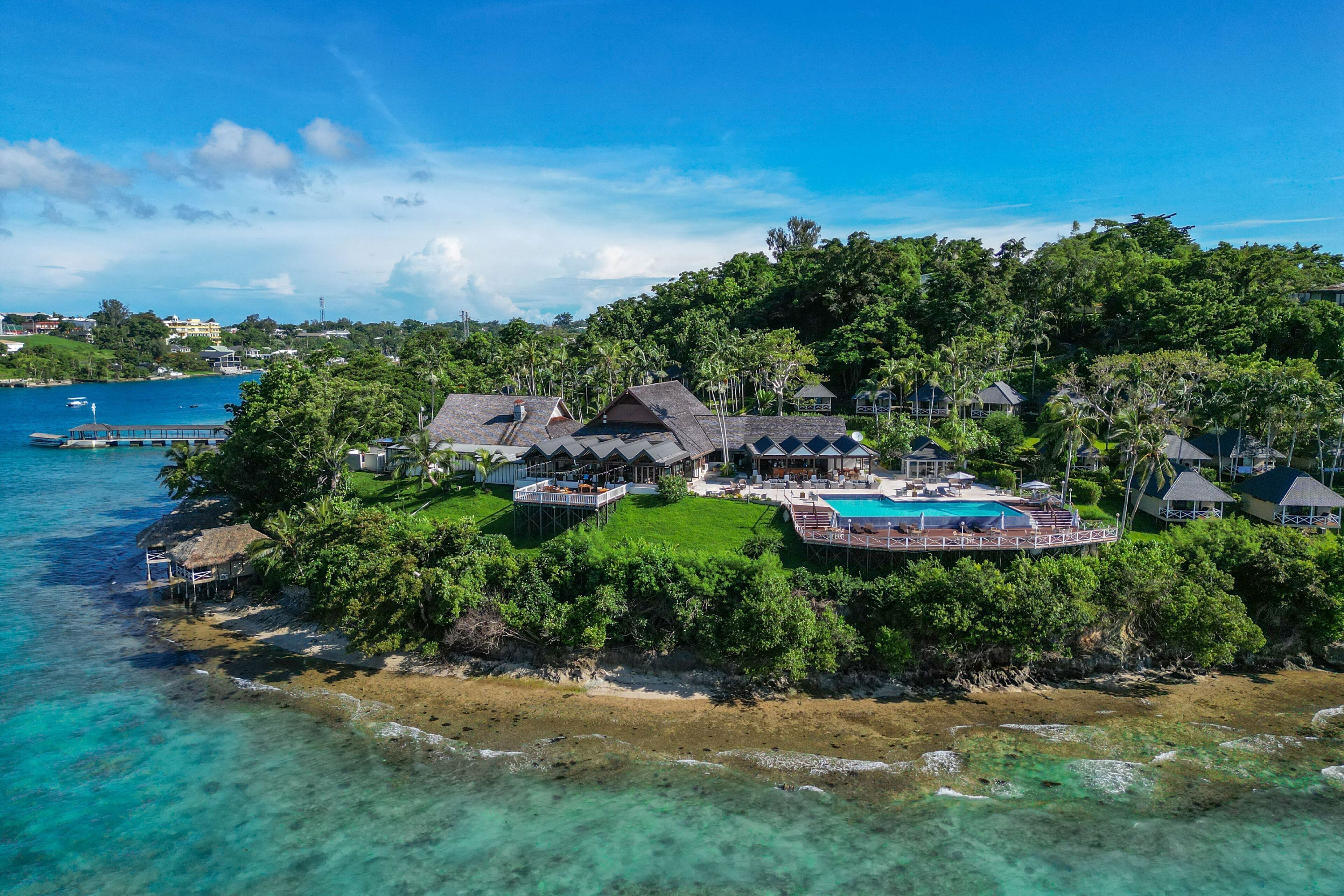An aerial view of a tropical resort with a large pool, multiple guest villas, and lush greenery.