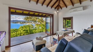 Una habitación con un amplio ventanal corredera que da a un balcón con vistas al mar y a las montañas. Hay dos sofás y una mesita con mantel.