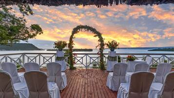 Una terraza de madera con sillas blancas y un arco floral, con vista a un cuerpo de agua al atardecer.