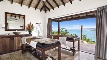 Una habitación con una cama, una camilla de masaje, un armario de madera, un espejo y vista al océano.