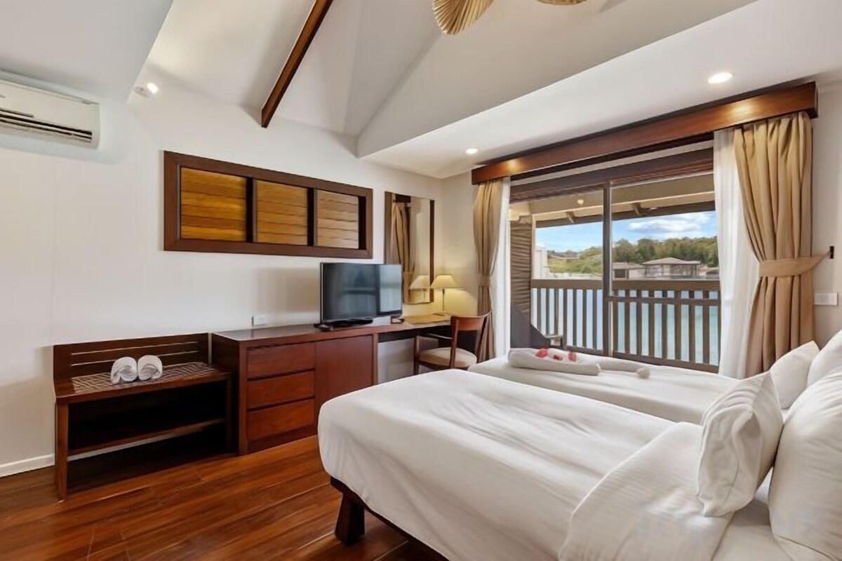 A hotel room with a bed, a desk with a television, a window with curtains, and a balcony with a view.