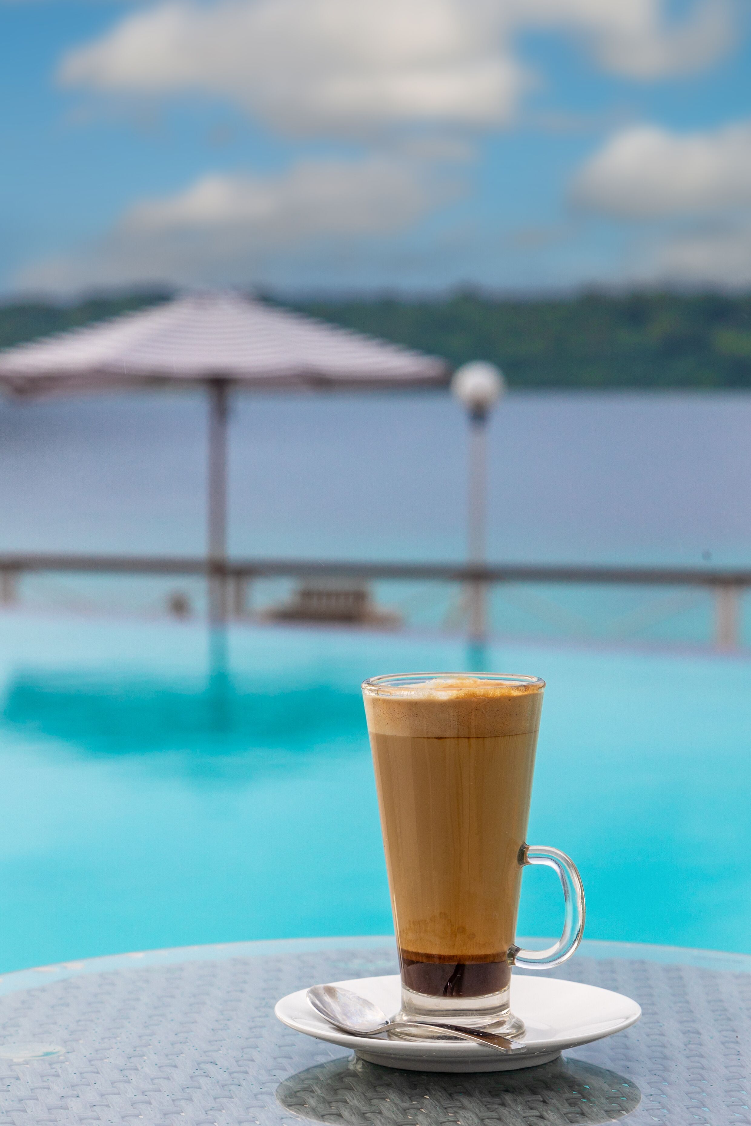 A glass of coffee on a table by a pool with a view of a distant island and a beach umbrella.