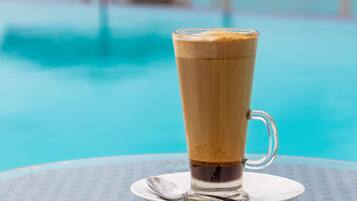 A glass of coffee on a table by a pool with a view of a distant island and a beach umbrella.