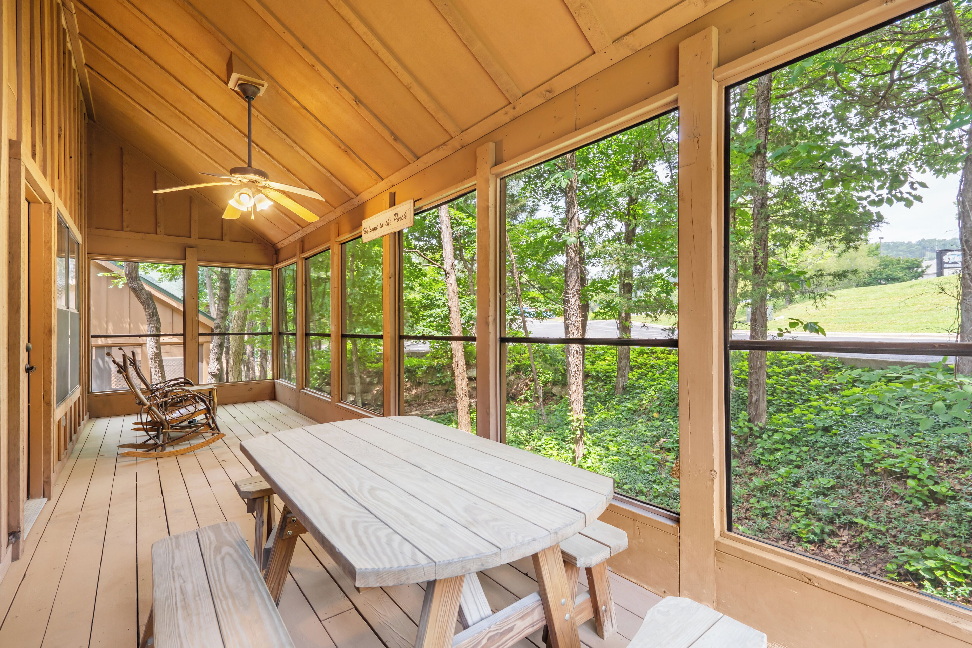 A wooden deck with a picnic table and chairs, large windows offering a view of trees and a grassy area.