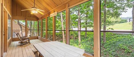 A wooden deck with a picnic table and chairs, large windows offering a view of trees and a grassy area.
