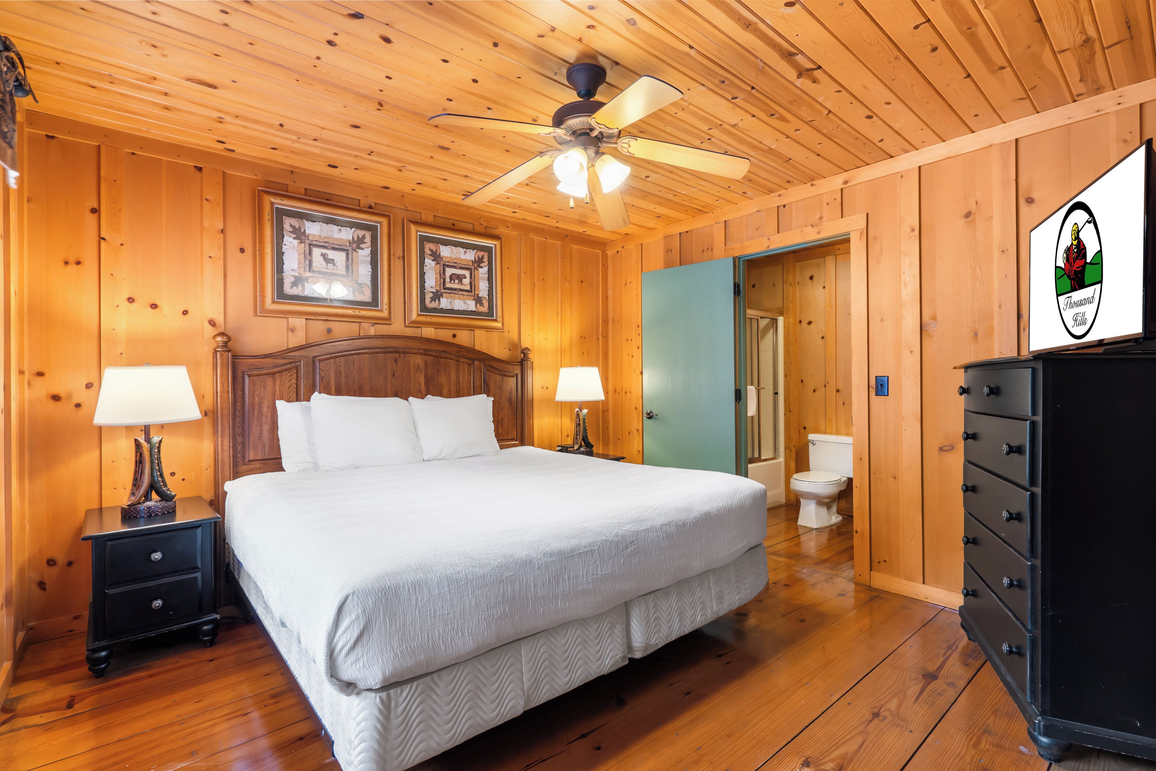 Camera da letto con rivestimento in legno, un letto, due comodini con lampade, una ventola a soffitto, un quadro incorniciato e un cartello sopra la porta.
