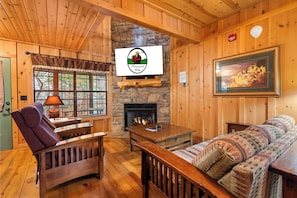 Interior acogedor de una cabaña con chimenea de piedra, muebles de madera, un televisor montado encima y un cuadro en la pared.