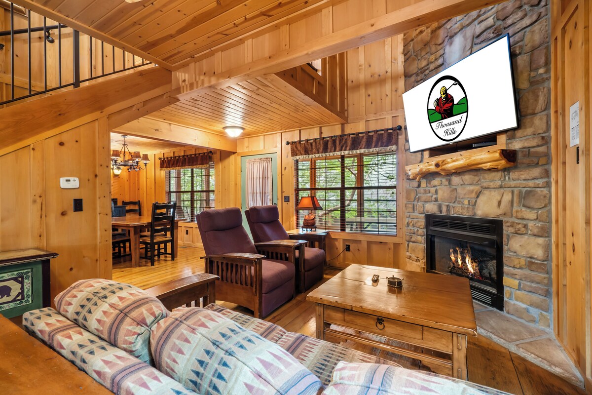 Interior acogedor de una cabaña de troncos con chimenea de piedra, muebles de madera y un televisor en la pared.