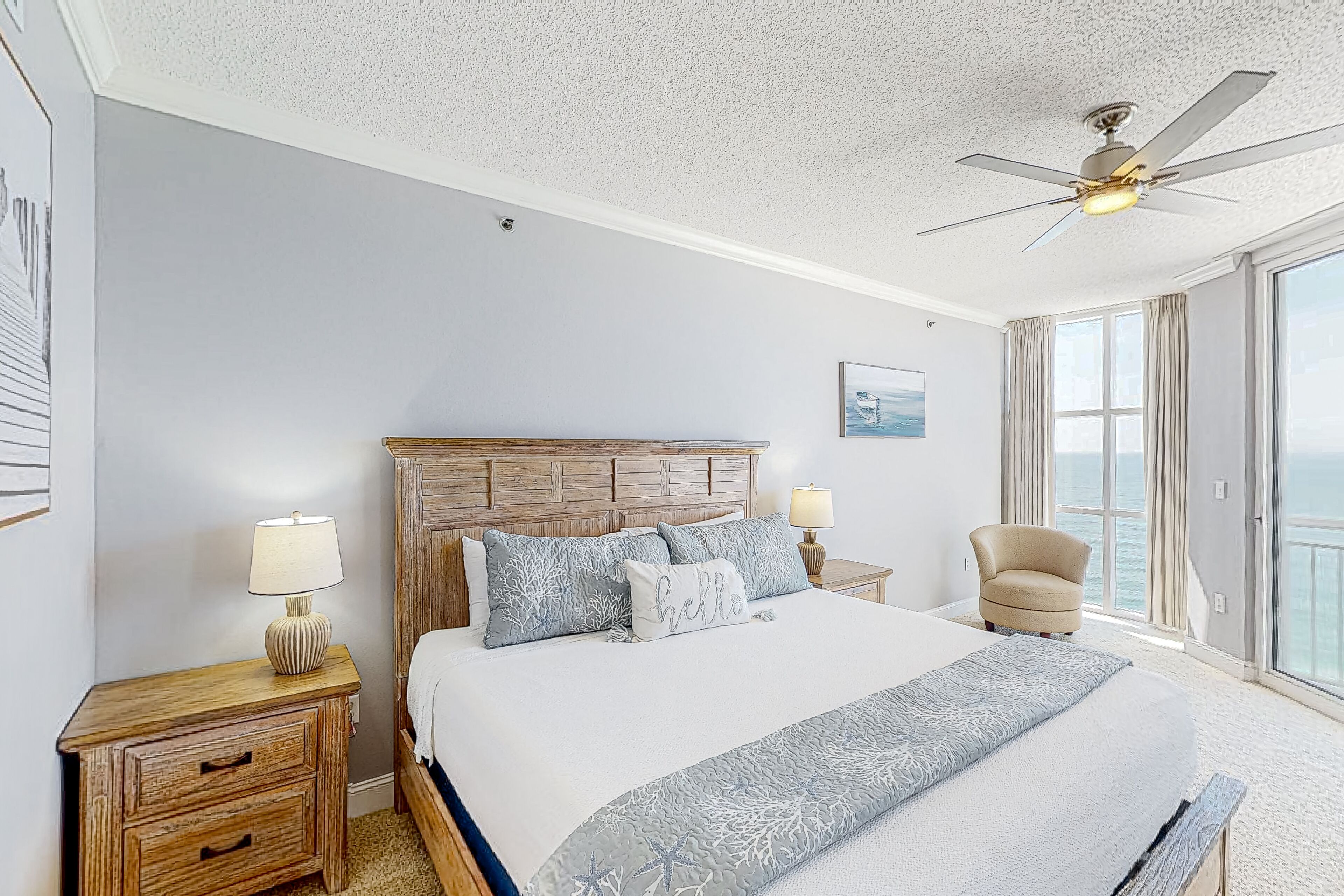 A bedroom with a large bed, wooden headboard, two bedside tables with lamps, a chair, and a view of the sea through large windows.