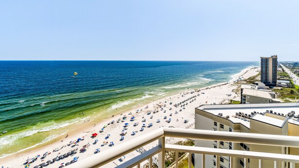 A beach view from a balcony with a clear sky, a sandy beach, and a tall building in the background. - Palacio Condominiums (Pensacola)