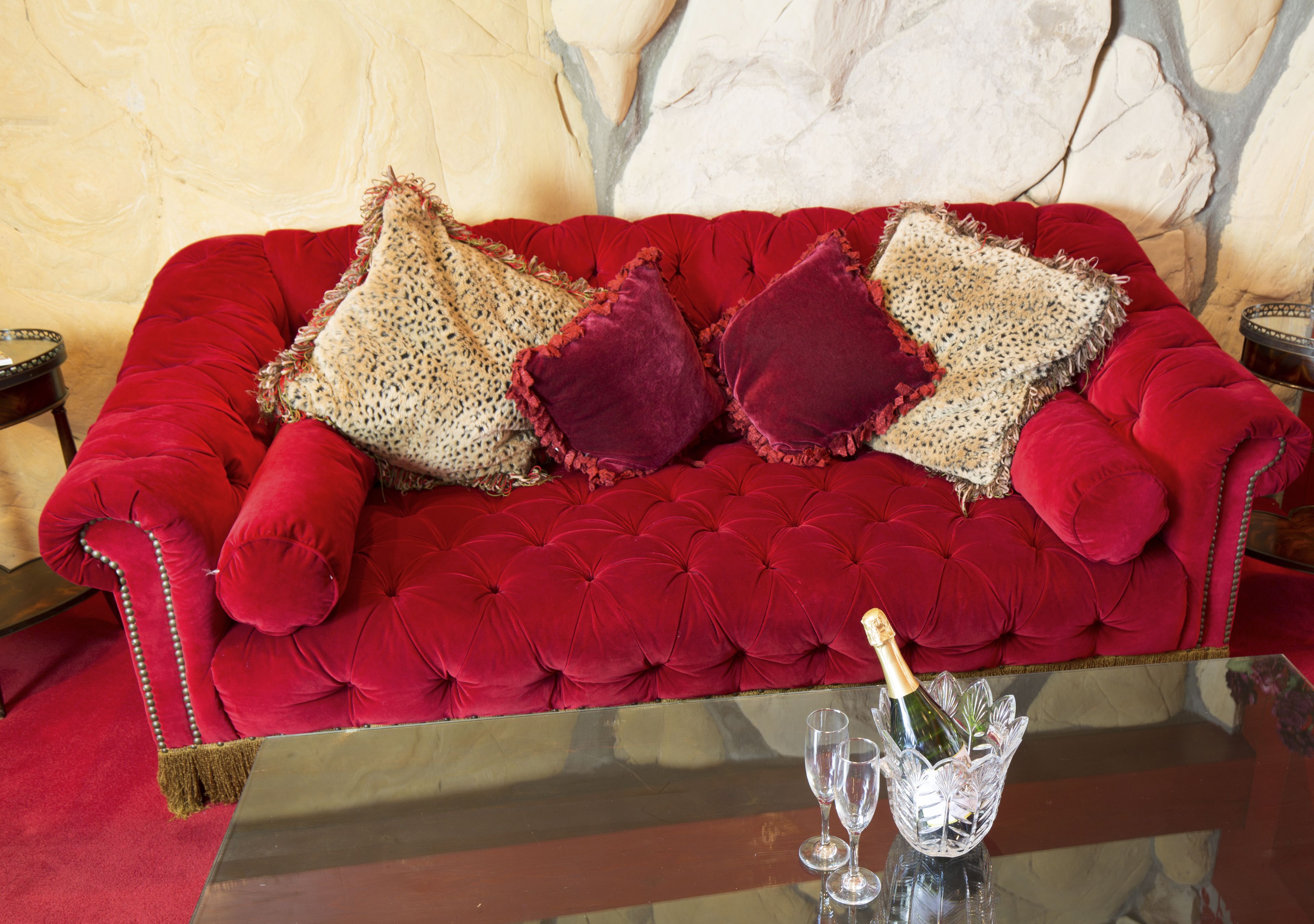 A red velvet sofa with decorative pillows and a glass table with a bottle and two glasses.