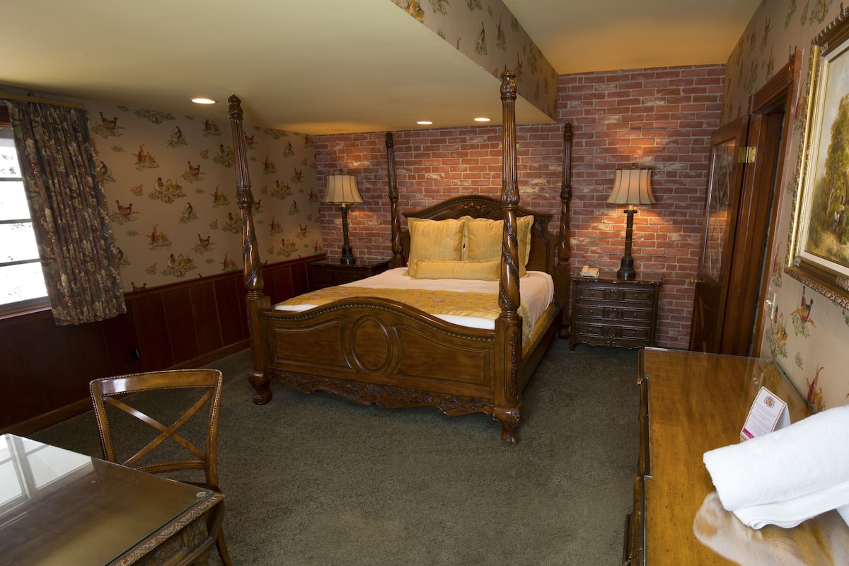 Ein Bett mit Baldachin, ein Holzkopfstück, ein Nachttisch mit Lampe, eine Holzkommode und ein Gemälde an der Wand.