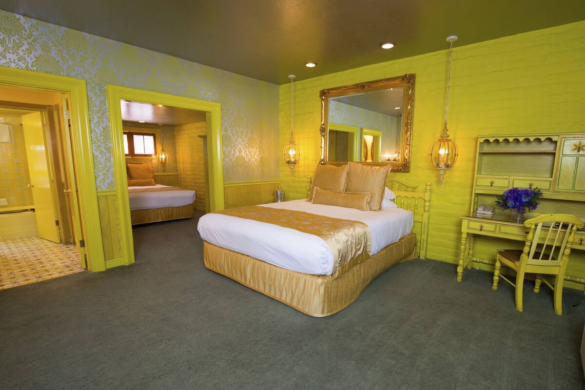 Ein Schlafzimmer mit einem großen Bett, einem Schreibtisch und einem Stuhl. Über dem Schreibtisch hängt ein Spiegel und eine Tür führt in einen anderen Raum.