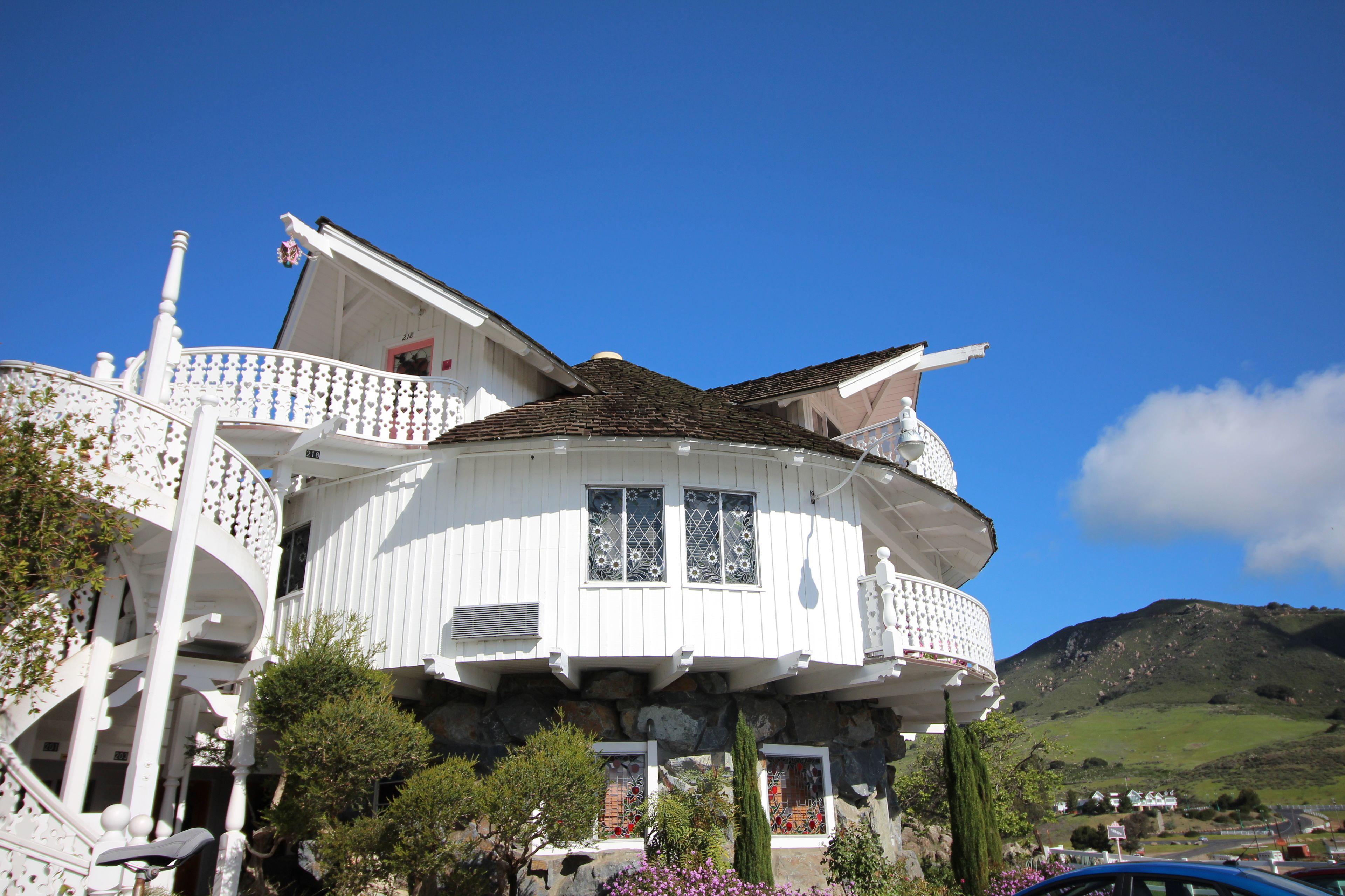 A white, multi-story house with a balcony, surrounded by greenery and a mountain in the background.