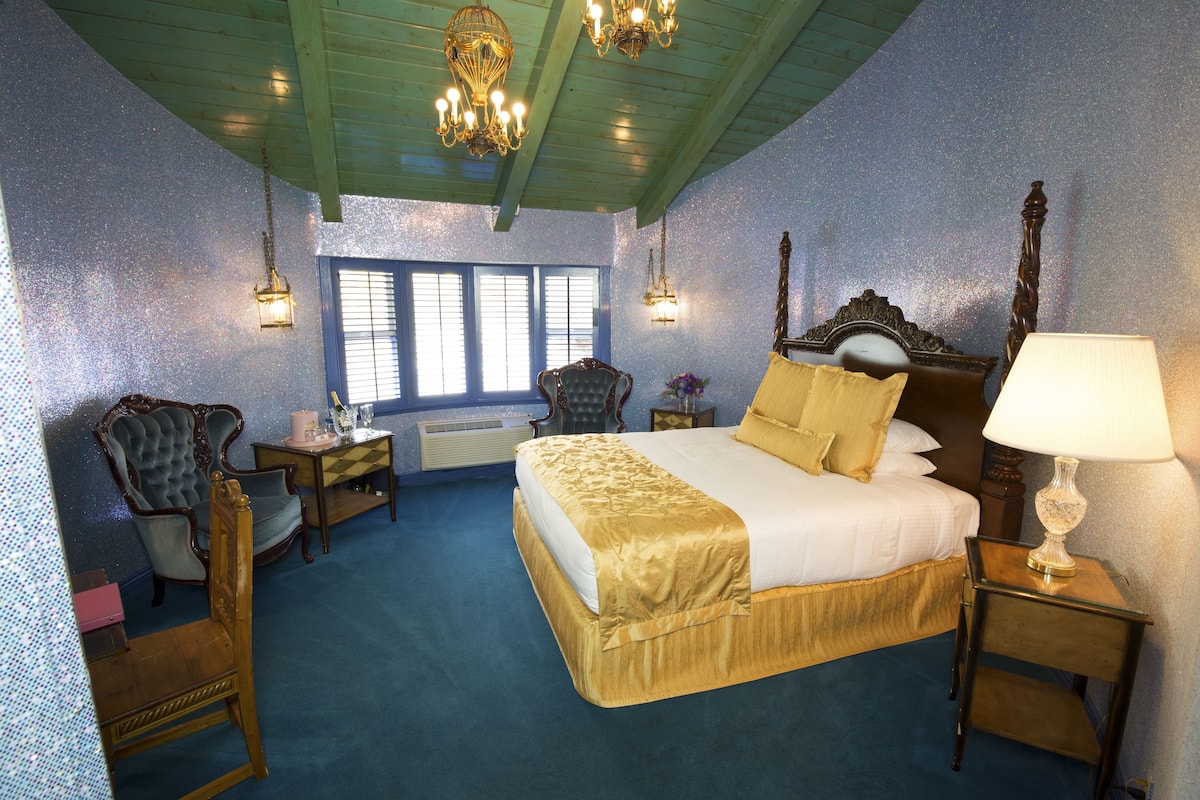 Ein Schlafzimmer mit einem großen Bett, zwei Stühlen, einem Nachttisch mit Lampe und einem Fenster mit Jalousien.