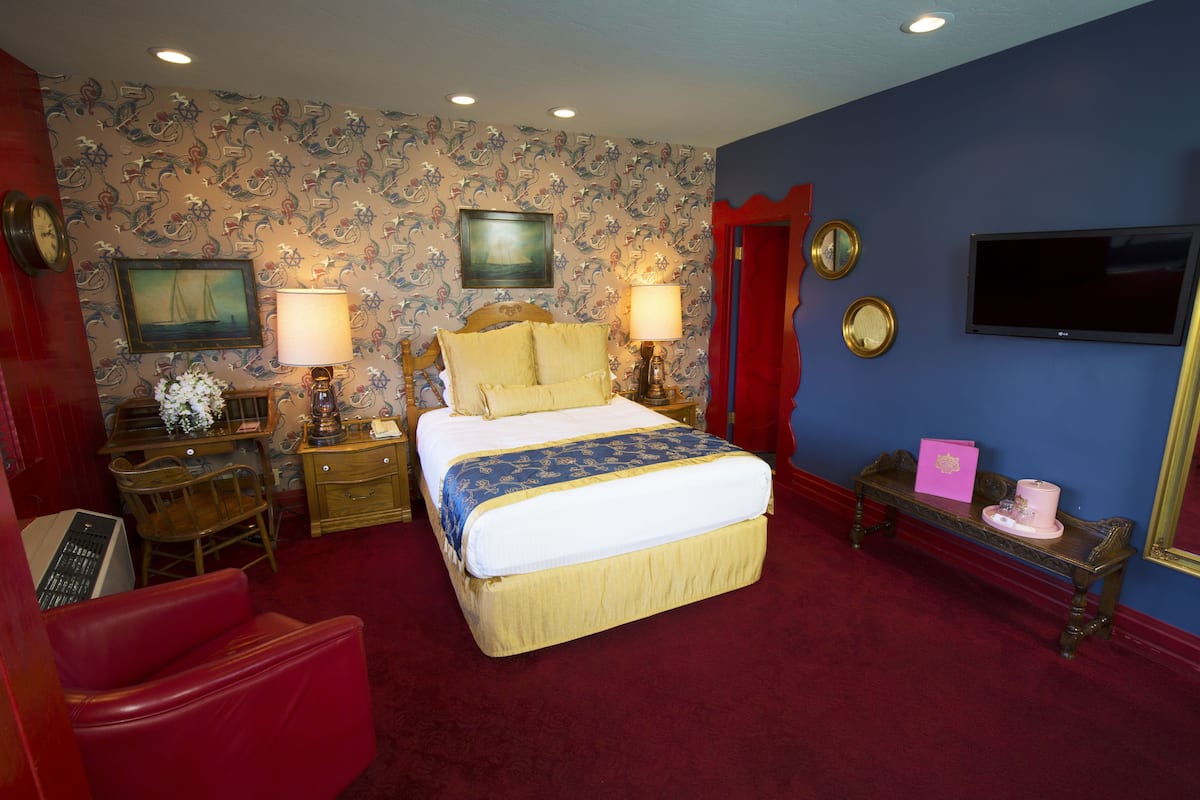 Ein Hotelzimmer mit einem großen Bett, zwei Nachttischen mit Lampen, einem roten Sessel, einem Schreibtisch mit Tastatur, einem Fernseher und einem Spiegel an der Wand.