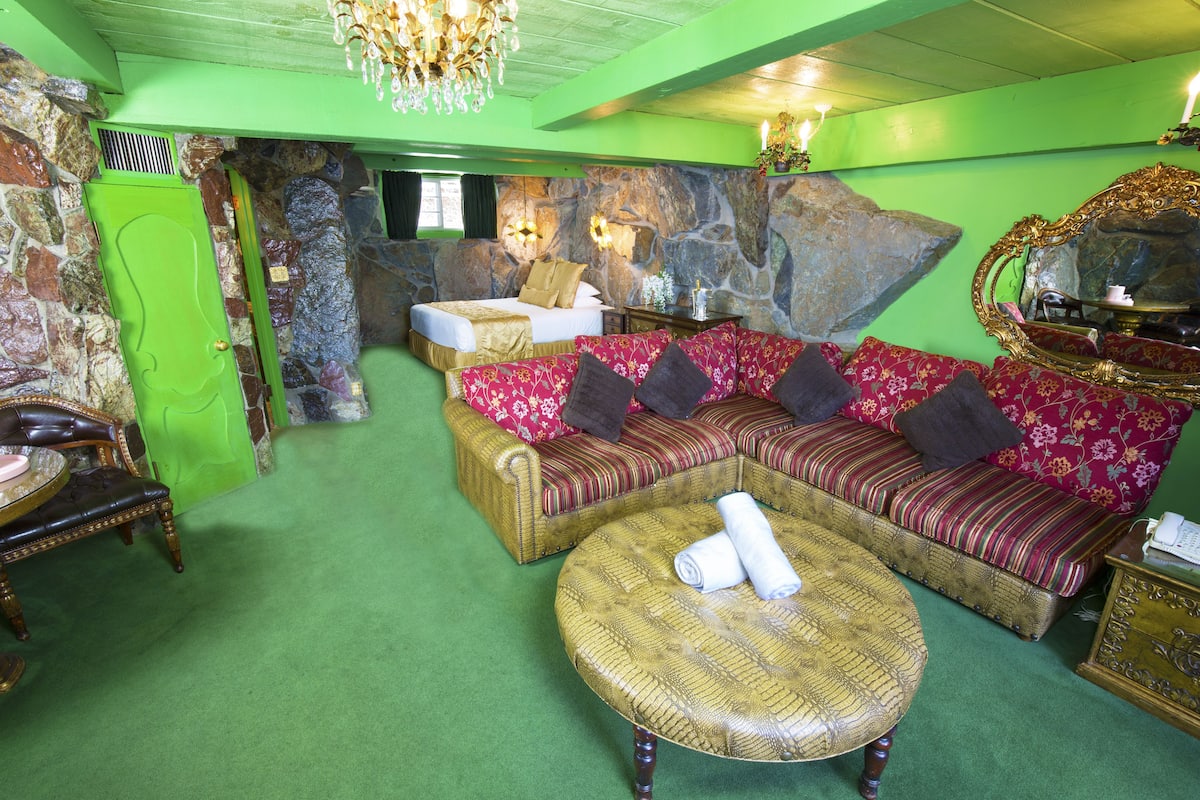 Ein Zimmer mit grünem Teppich, einer großen Sofagarnitur, einem runden Korbstuhl-Tisch und einer Steinwand.