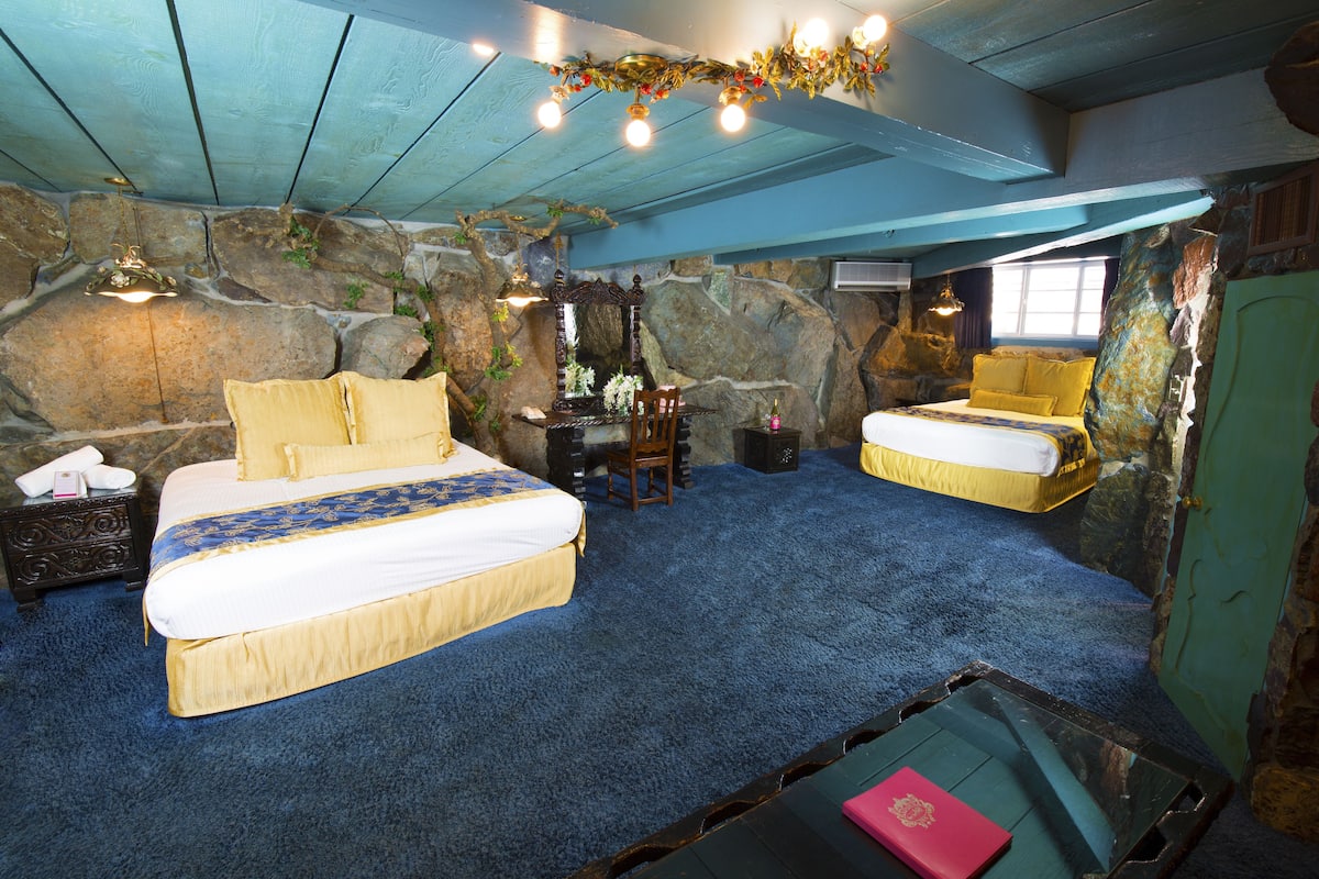 Ein Schlafzimmer mit einem großen Bett, einem kleineren Bett, einem Schreibtisch und einem Stuhl. Eine Steinwand und ein Fenster sind ebenfalls vorhanden.