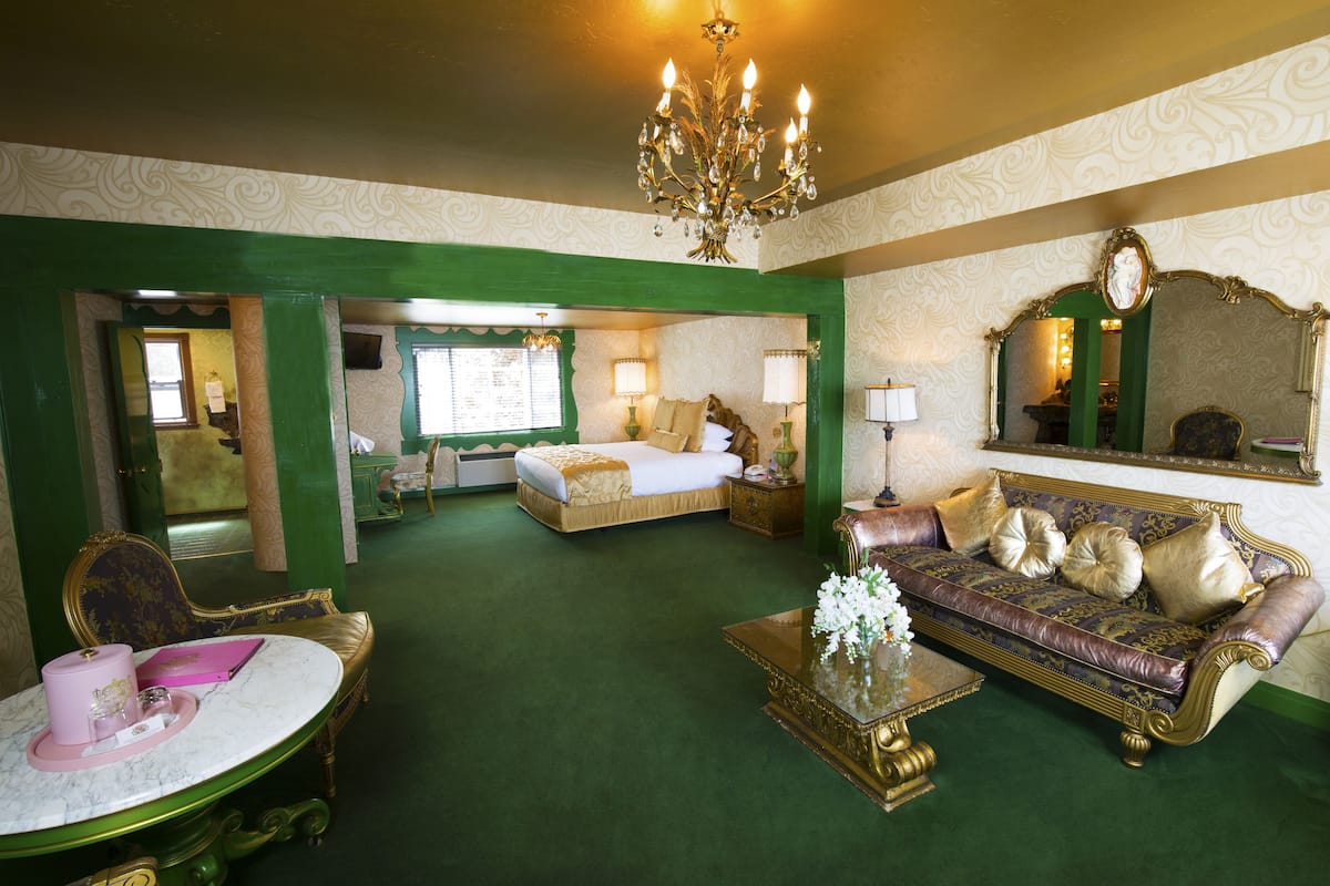 Ein luxuriöses Zimmer mit grüner und goldener Farbgestaltung, ausgestattet mit einem Bett, einer Couch, einem Tisch mit einer Vase und einem Spiegel.