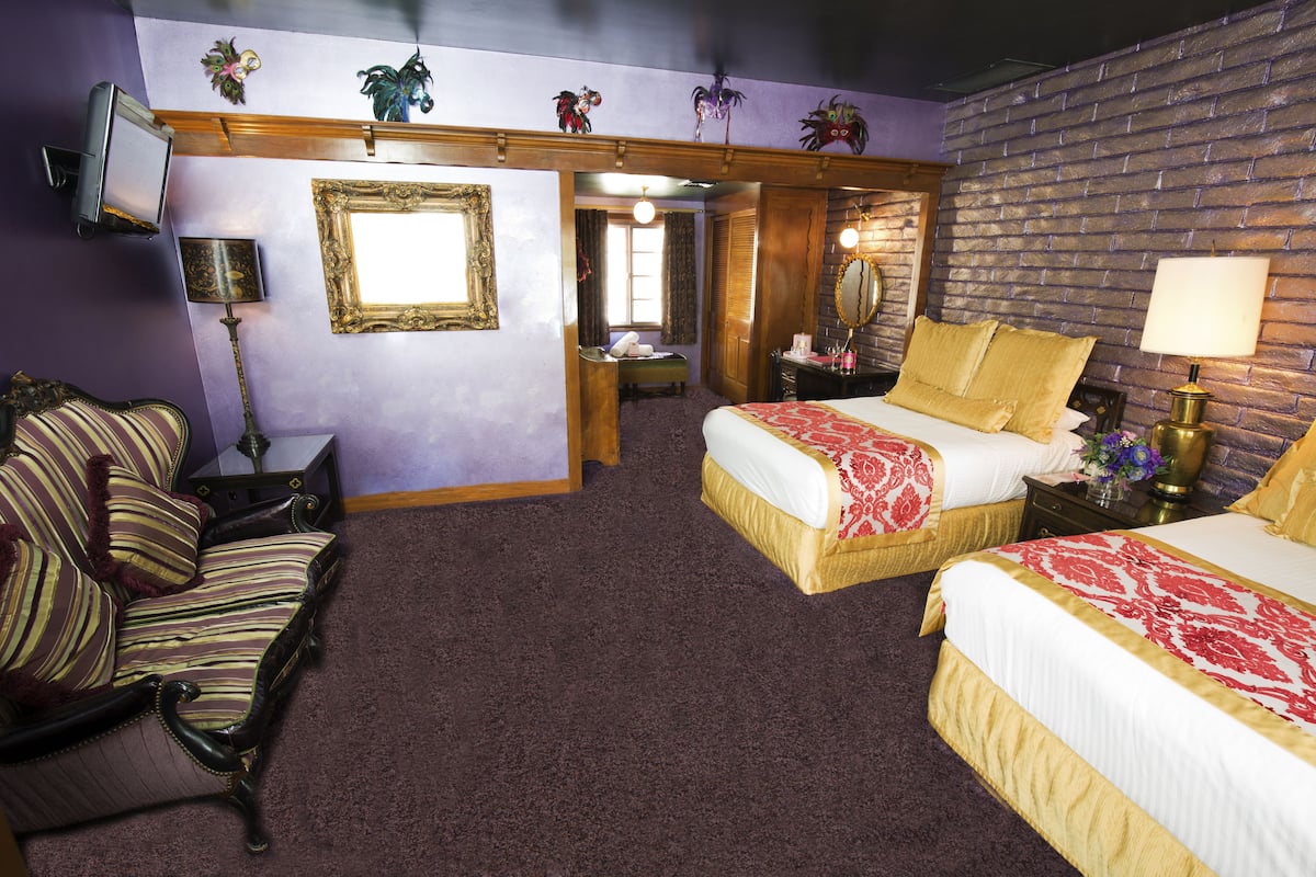 Ein Hotelzimmer mit zwei Betten, einem Sessel, einem Schreibtisch, einem Spiegel und einem Fernseher.