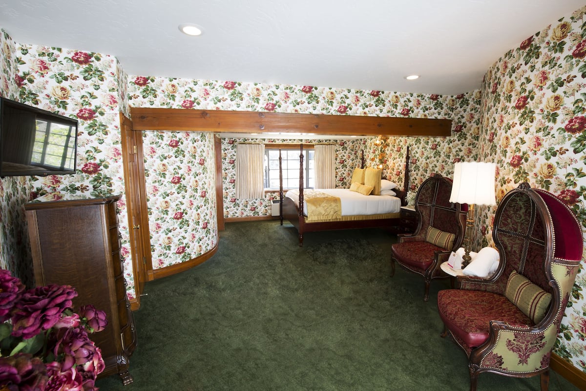 Ein Schlafzimmer mit floraler Tapete, einem Holzbettgestell, einem grünen Teppich und zwei Polstersesseln.
