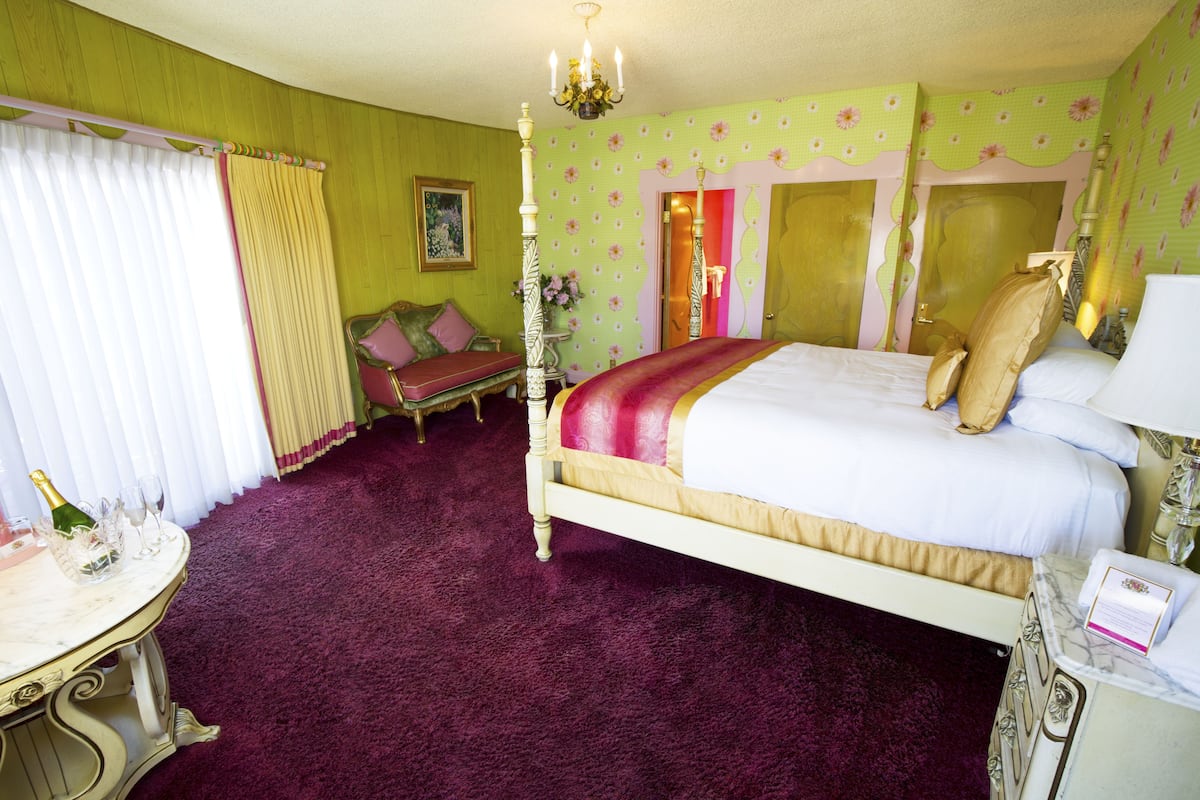 Ein Schlafzimmer mit einem Himmelbett, einem Schminktisch mit Spiegel, einem kleinen Tisch mit Flasche und Gläsern sowie einer Tür zu einem weiteren Raum.